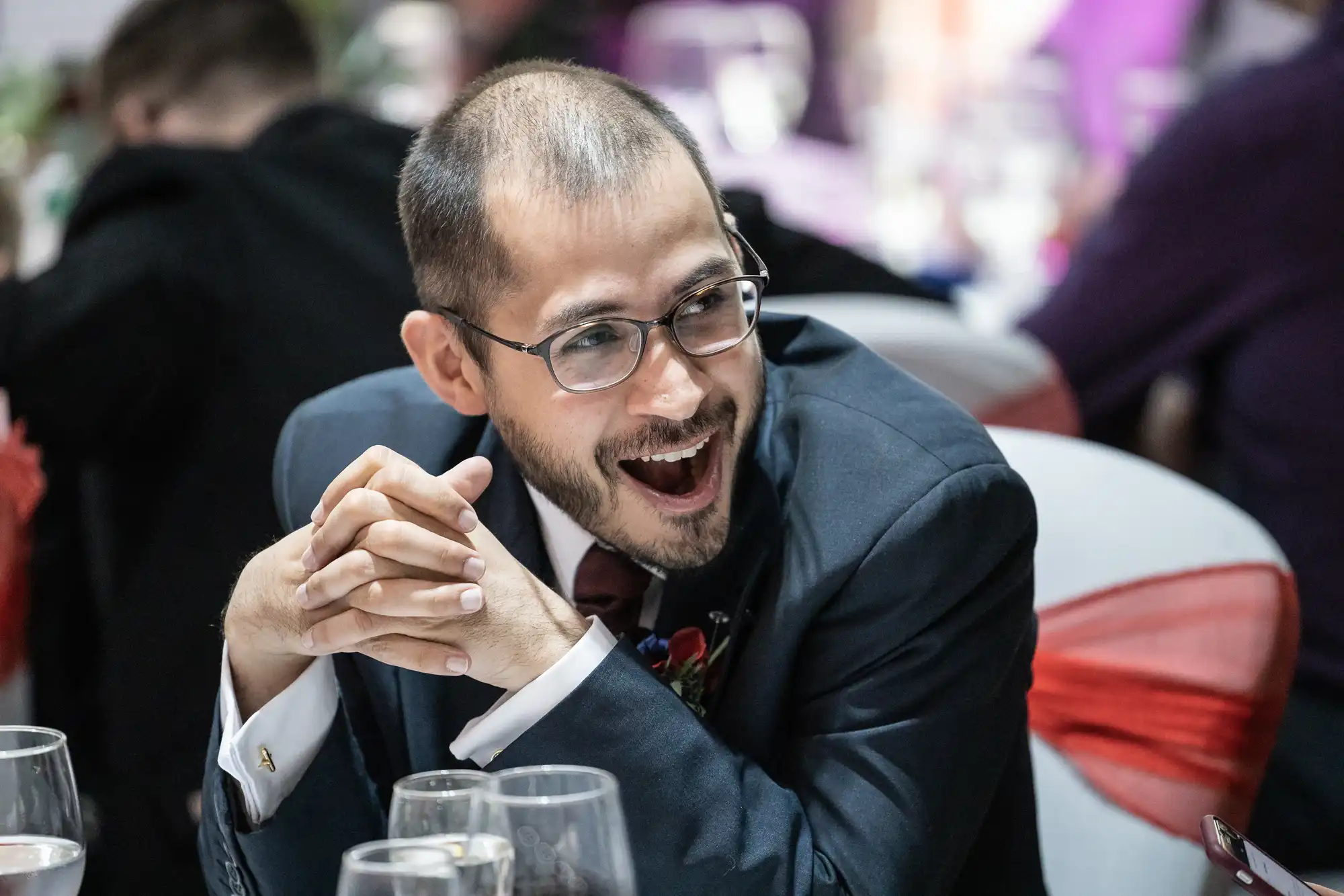 A man with glasses and a beard, dressed in a suit and tie, sits at a formal event table smiling widely with his mouth open, hands clasped together. Glasses and blurred people are visible in the background.