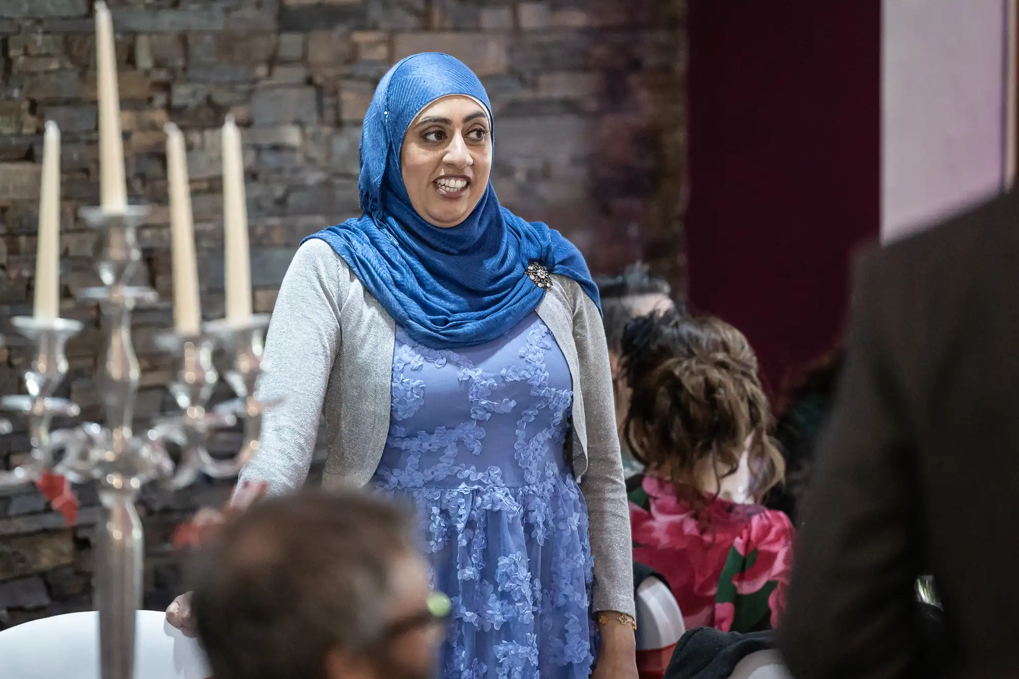 A woman in a blue hijab and lace dress with a grey cardigan stands smiling at an indoor event. People are seated around her, and a decorative candelabrum is visible to the left. The background features a stone wall.