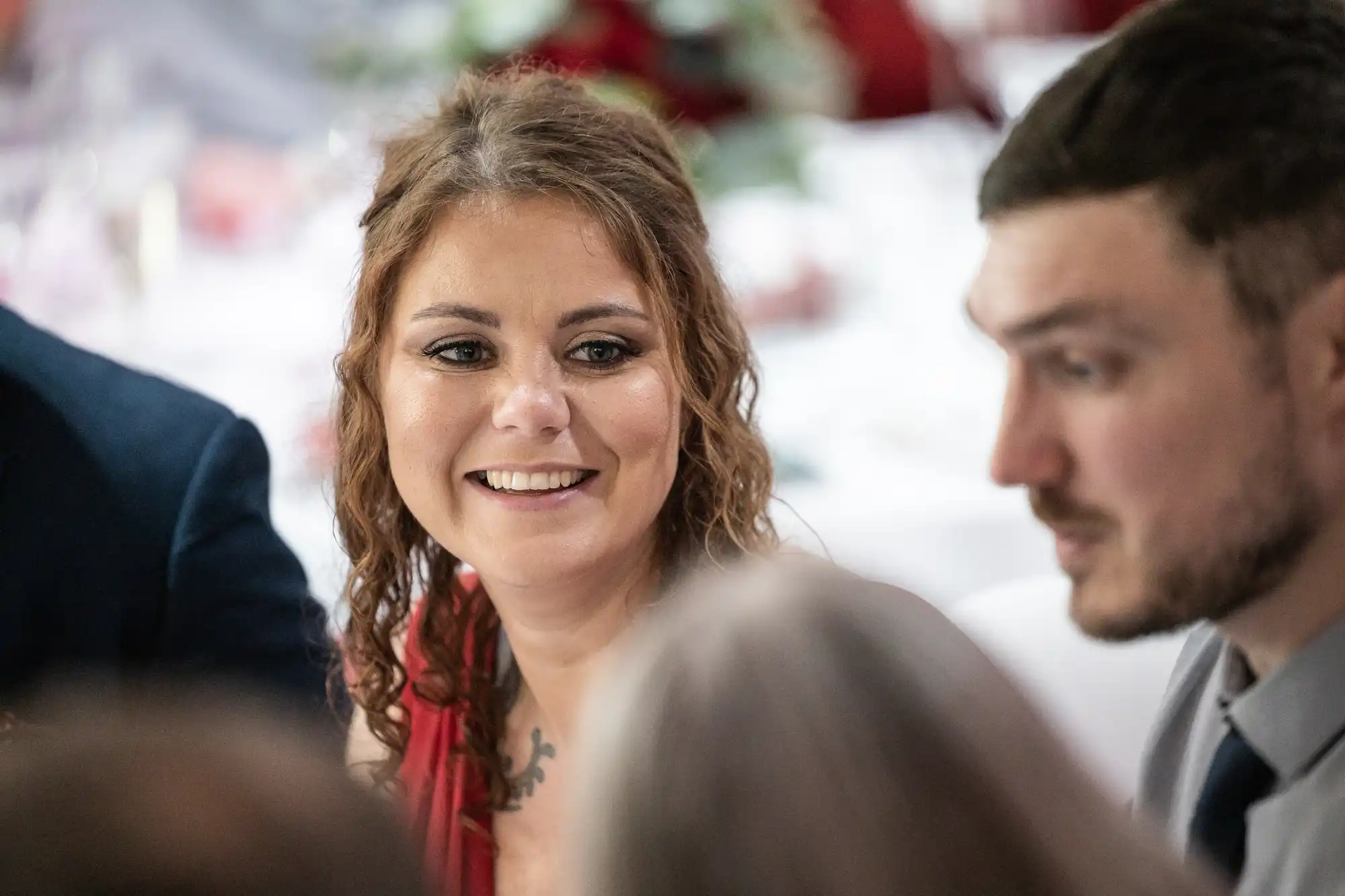 A woman with wavy brown hair and a red dress smiles whilst sitting at a table, surrounded by people. The background is softly blurred, suggesting a formal or celebratory event, such as a wedding or dinner party.