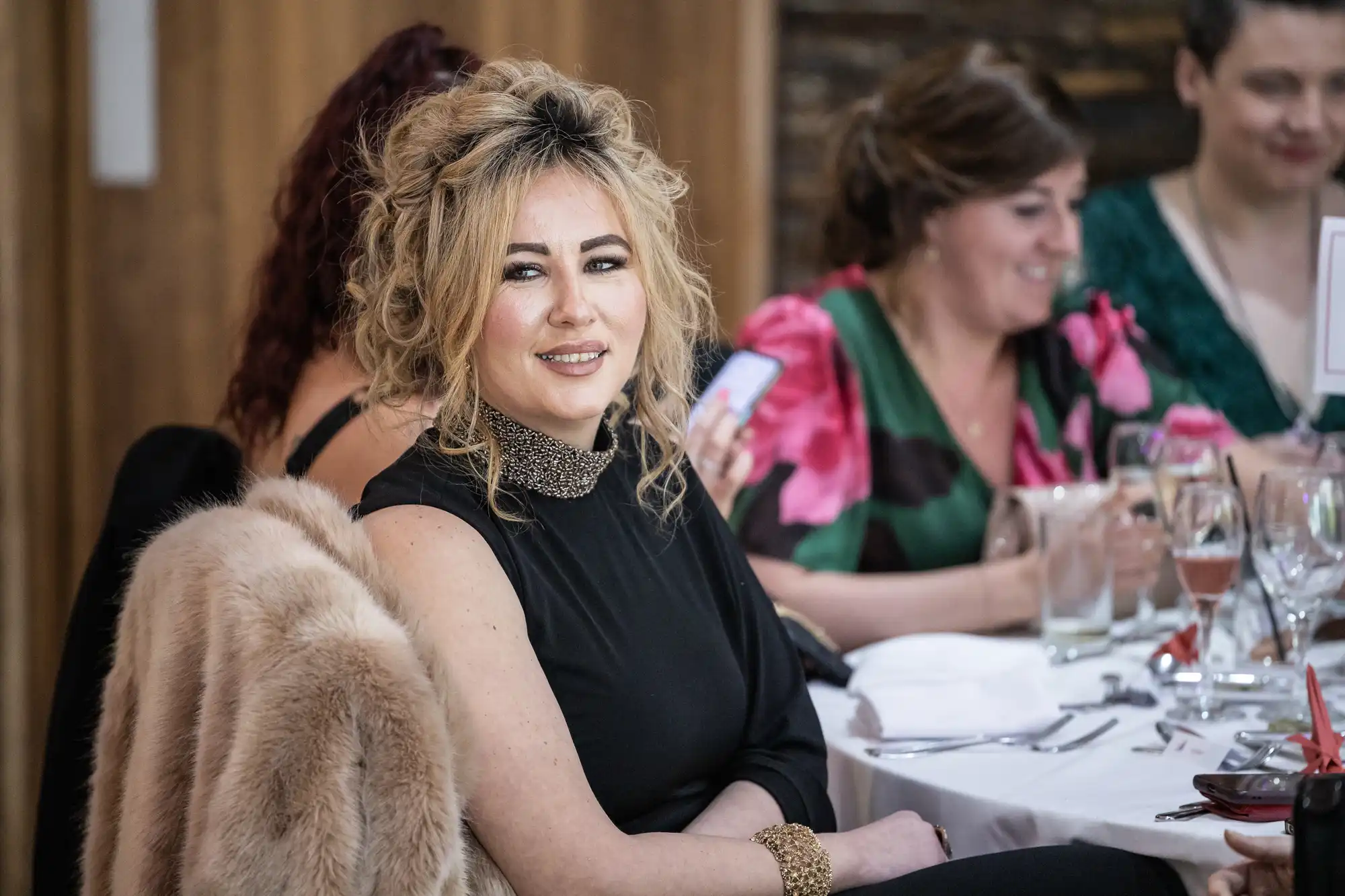 A woman with curly blonde hair in a black dress and gold accessories sits at a round table set for dining, with a fur coat draped over her chair. Other women are seated around her, engaged in conversation. The setting appears to be a formal event or celebration.