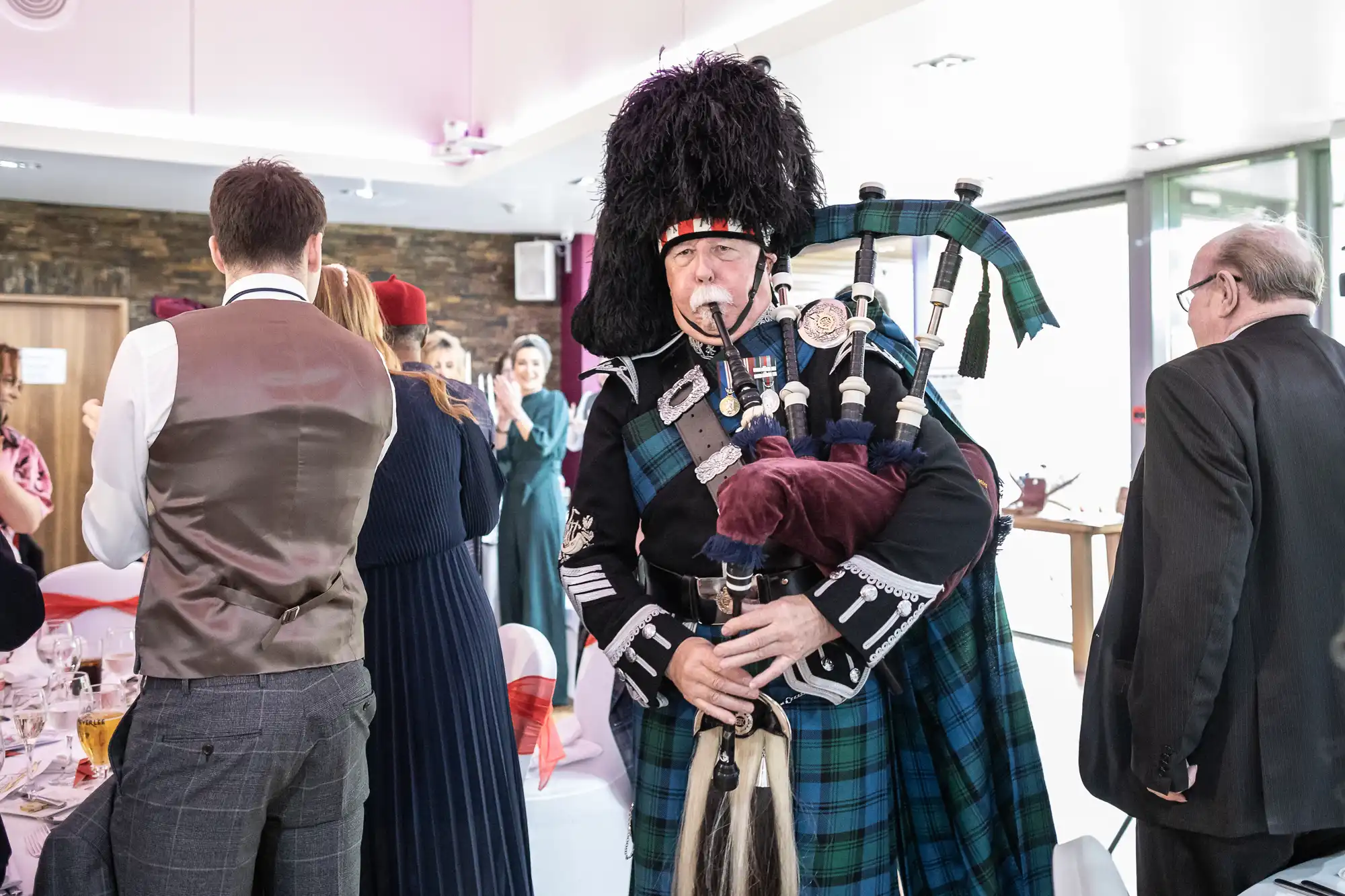 A man in traditional Scottish attire plays bagpipes at an indoor event. He wears a blue tartan kilt, black jacket, and a tall feathered hat. People in formal clothing stand and watch, with tables set for a meal in the bright, modern room.