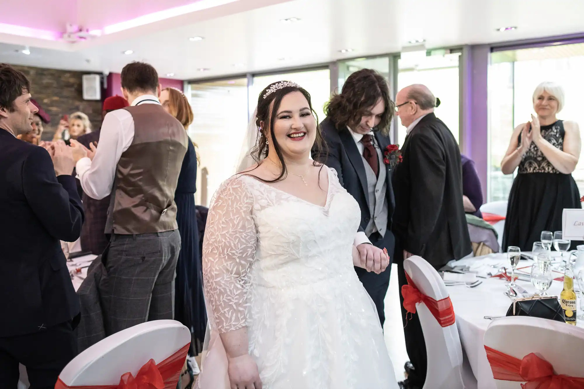 A smiling bride in a white lace wedding dress stands in a bright reception room. Guests in formal attire clap and mingle around decorated tables with red ribbon accents, whilst natural light streams through large windows in the background.