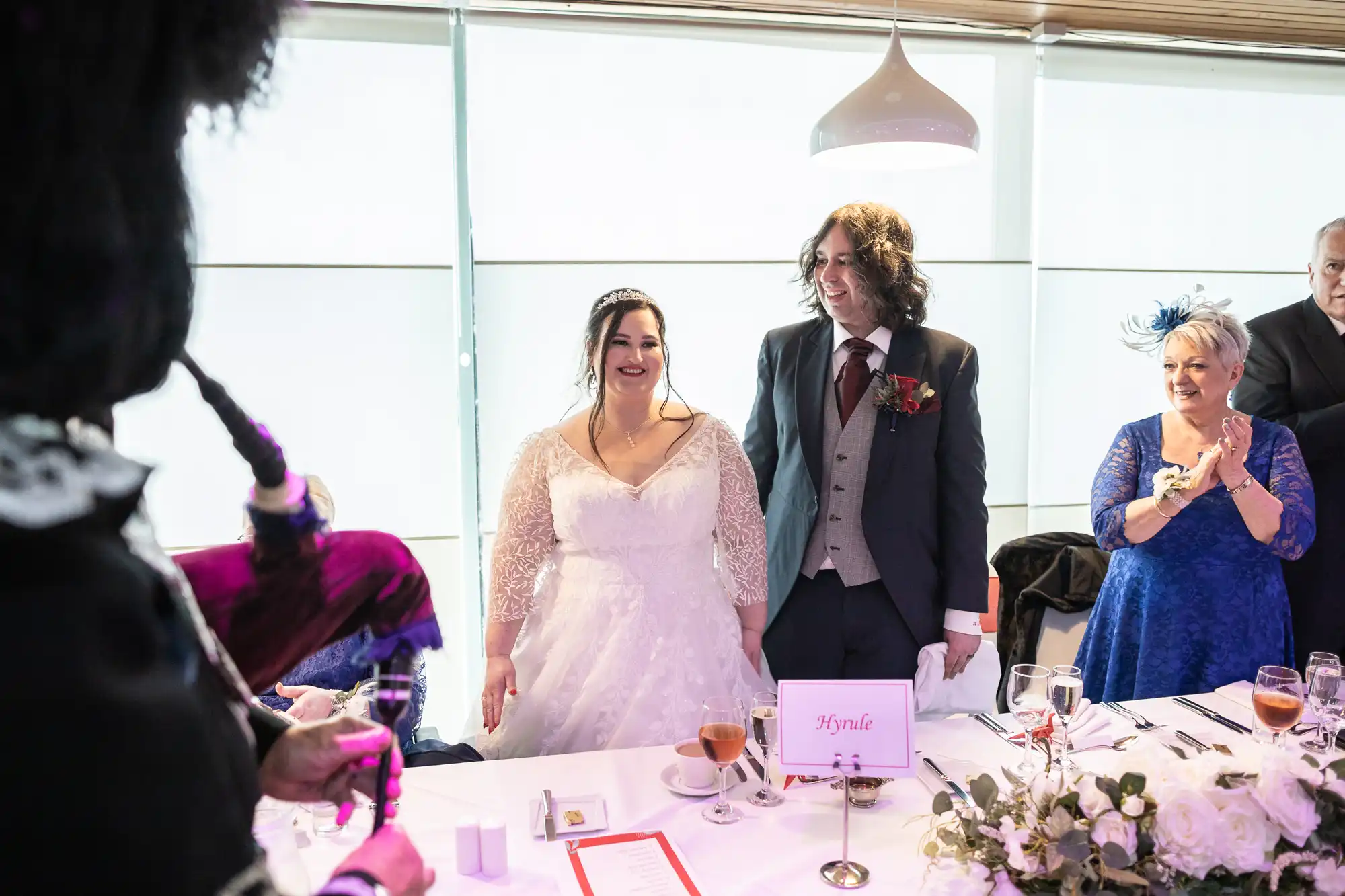 A bride in a lace wedding dress and groom in a suit stand smiling behind a table with flowers, drinks, and a "Krysta" place card. Guests in formal attire are around them, and a bagpiper is playing in the left foreground. Bright window light fills the room.