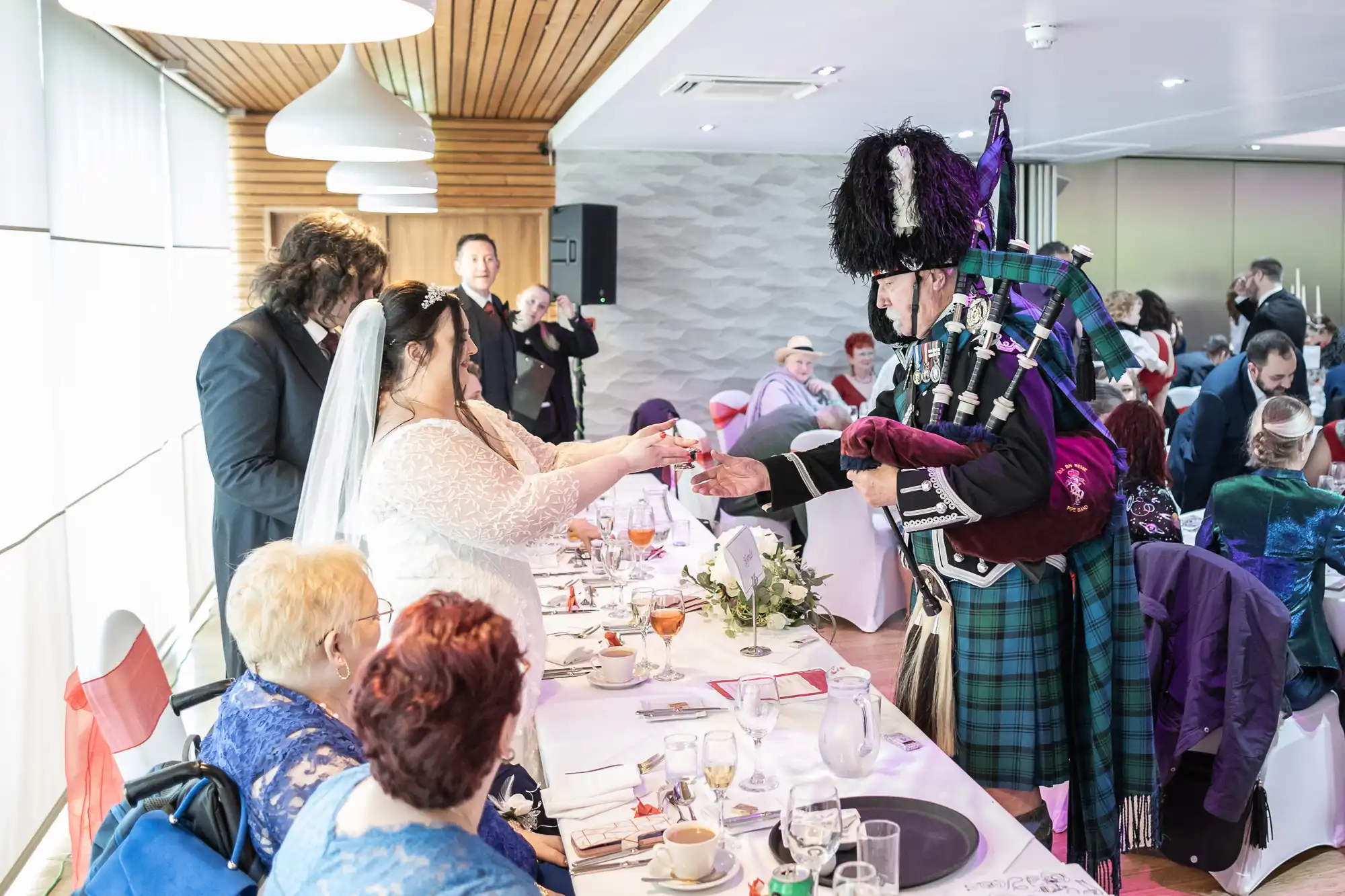 A wedding reception scene: a bride in a white dress shakes hands with a man in traditional Scottish attire and bagpipes. Guests sit at a decorated table with food and drinks. The room is bright, with wooden and modern décor elements visible.