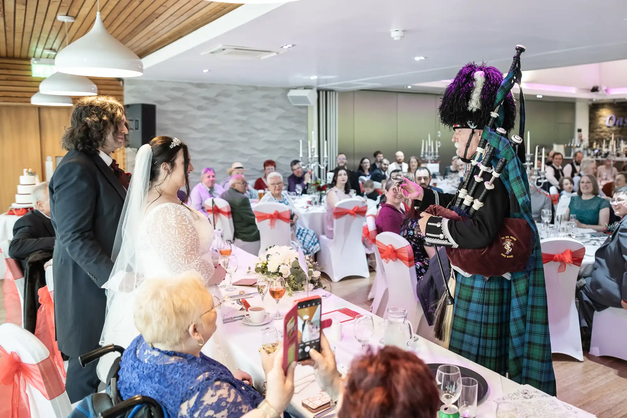 A bride and groom in wedding attire stand smiling before a bagpiper in traditional Scottish dress, performing at a lively reception. Guests seated at decorated tables watch, some taking photos; the room is bright with festive decor and red chair sashes.