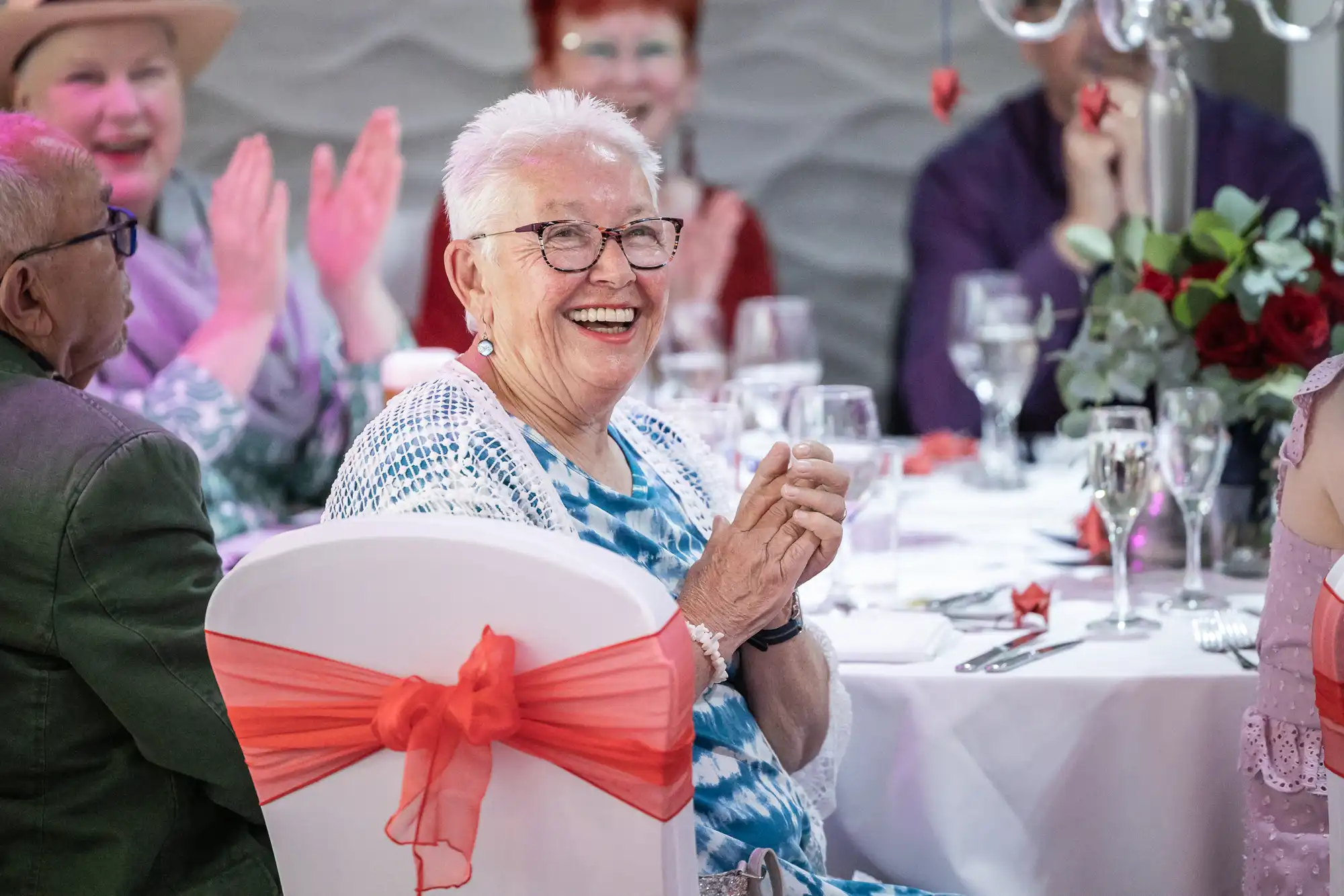 An older woman with short white hair and glasses smiles joyfully at a decorated table during a celebration. She wears a blue patterned top; orange ribbon decorates her chair. Several others sit nearby, clapping and laughing, with floral centrepieces and glassware visible.