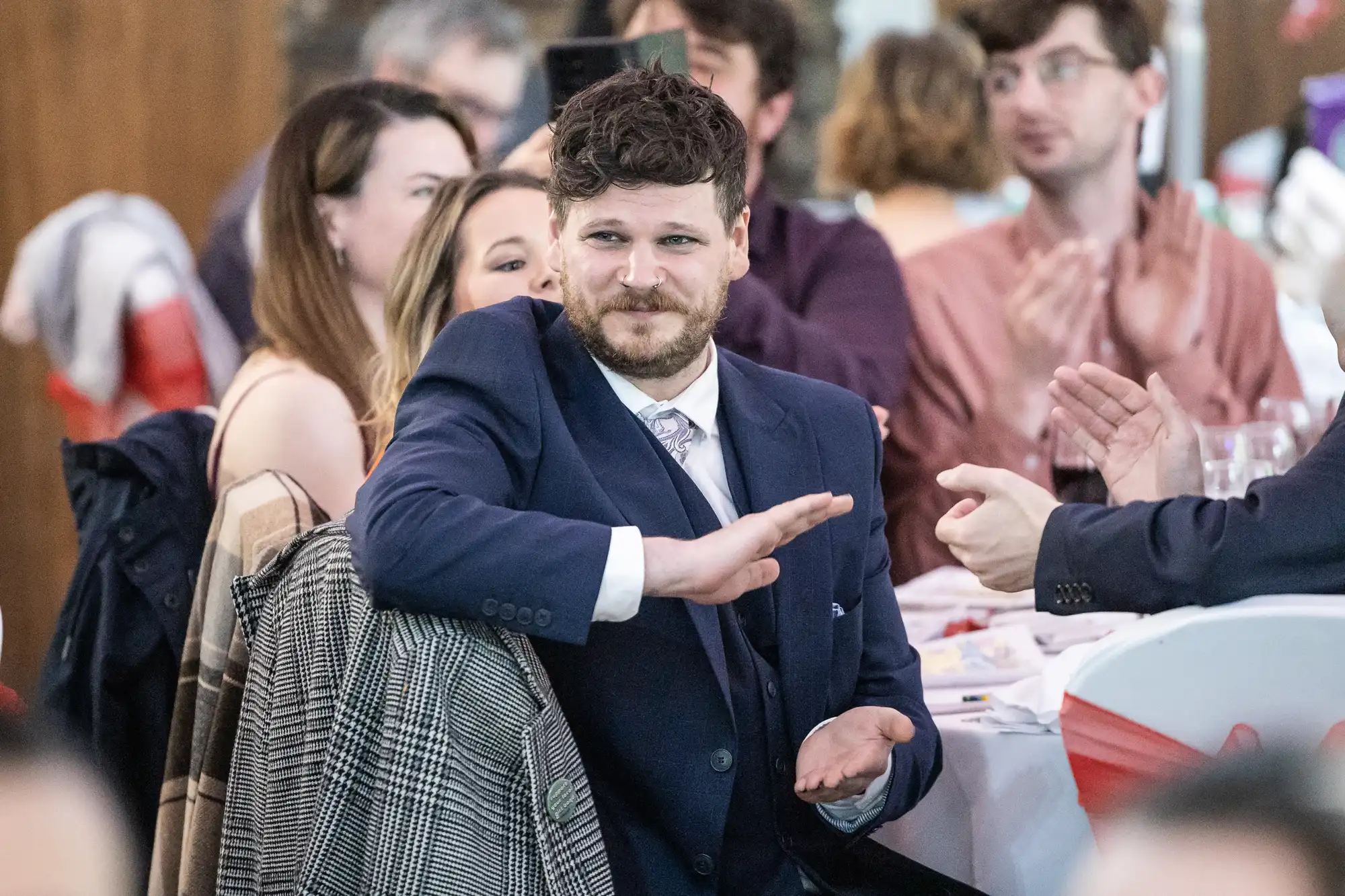 A man with curly hair and a beard, wearing a navy suit and tie, sits at a table applauding at a formal event. He is surrounded by other seated guests, some clapping, with white tablecloths and red decorations visible. The atmosphere is lively and sociable.
