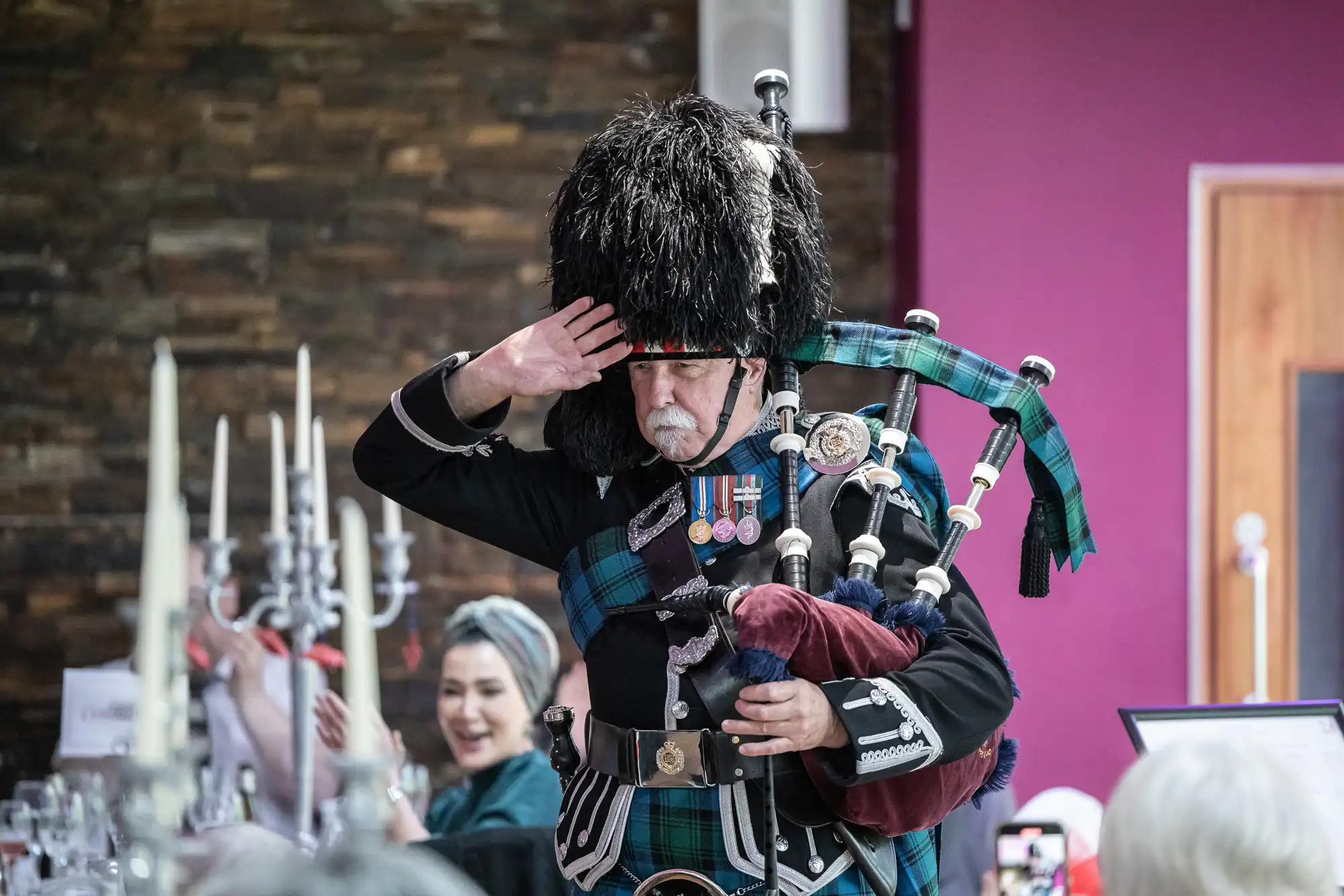 An older man in ceremonial Scottish attire, including a feathered hat and tartan, plays the bagpipes and salutes. He stands indoors before a seated audience, with candelabra and stone and pink walls visible in the background.