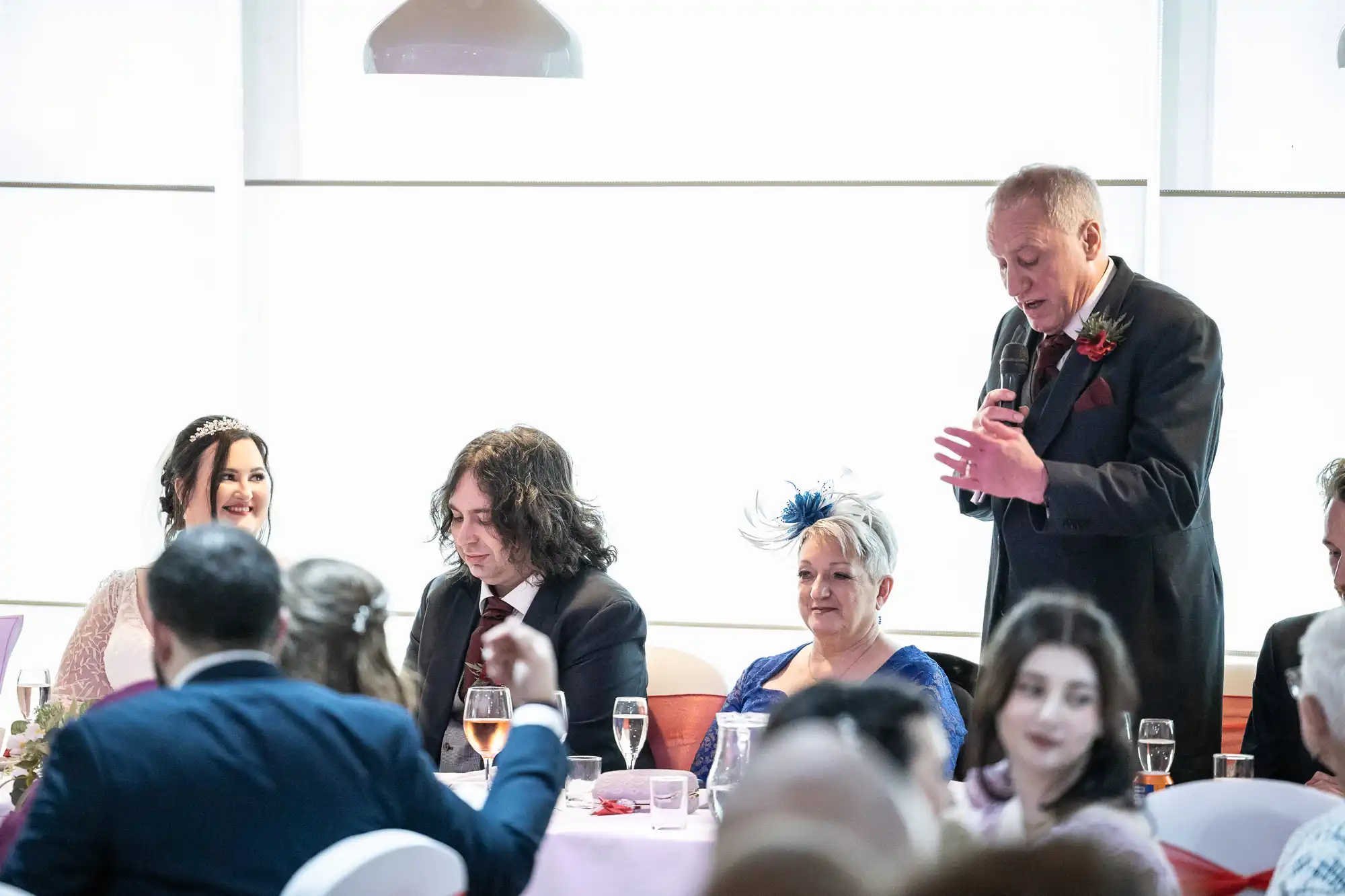 A man stands and speaks into a microphone at a wedding reception table. Guests, including a smiling bride in a tiara, a man with long dark hair, and a woman in a blue dress with a fascinator, listen. Wine glasses and plates are on the pink-covered table.