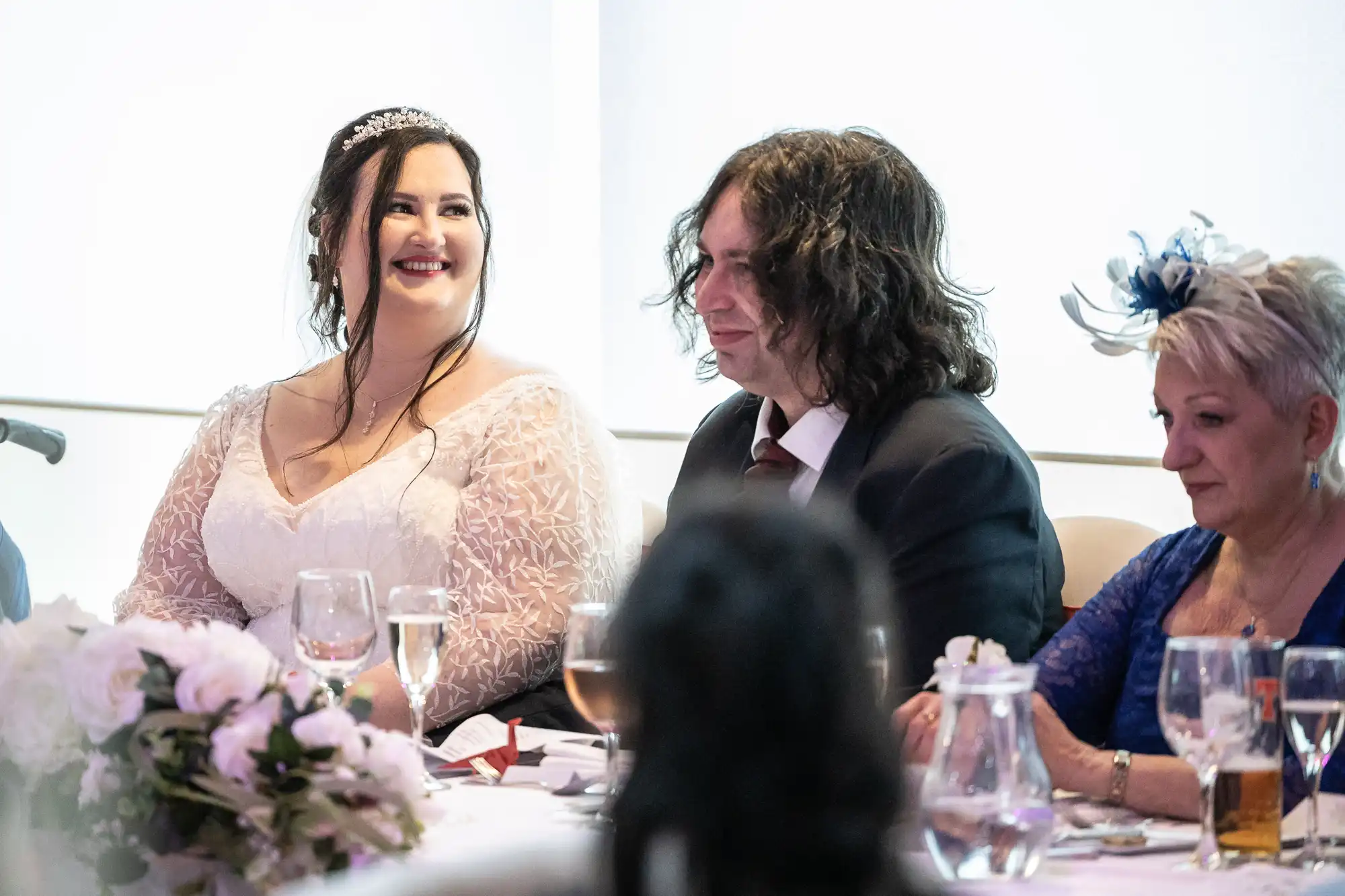 A bride in a white dress smiles beside a man in a suit and tie, with a woman in a blue dress and fascinator seated next to them. They sit at a decorated table with flowers, glasses, and a jug, during what appears to be a wedding reception.