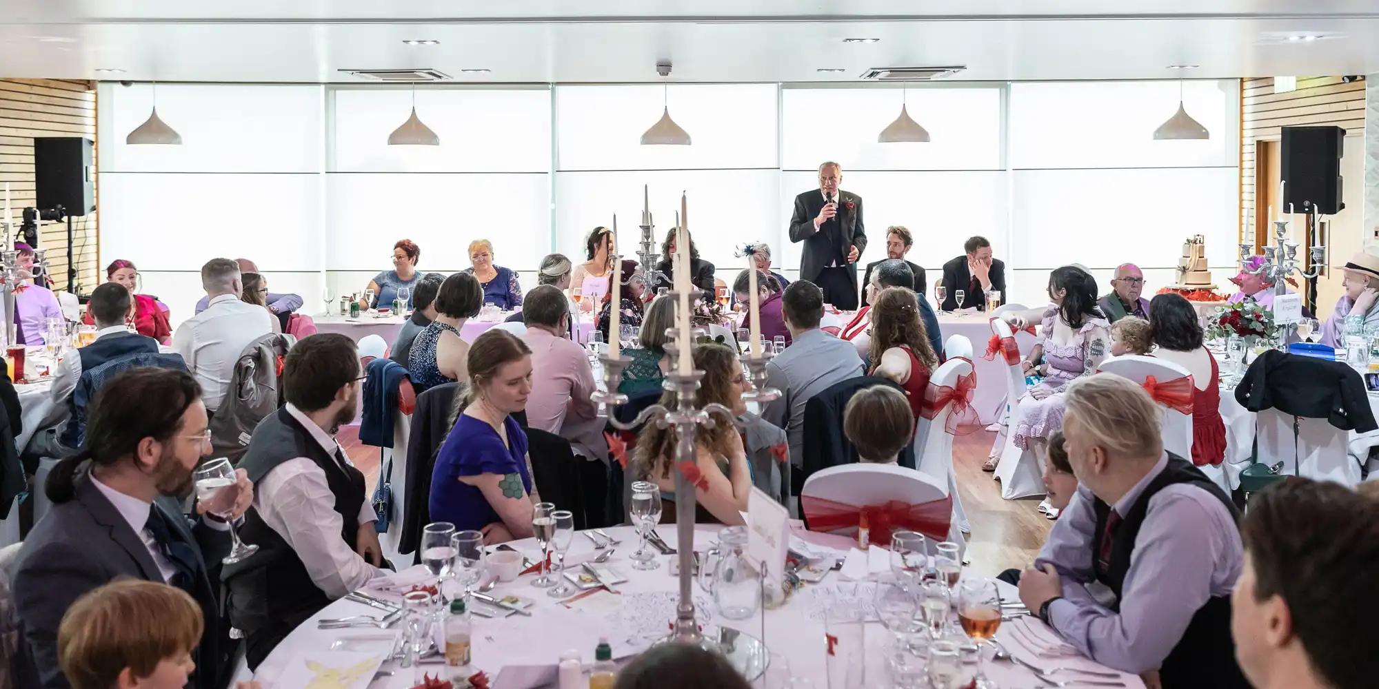 A large group of people sit at round tables decorated with white cloths and red sashes, watching a man in a suit give a speech at the front of a bright, modern banquet hall with large windows and hanging lights.