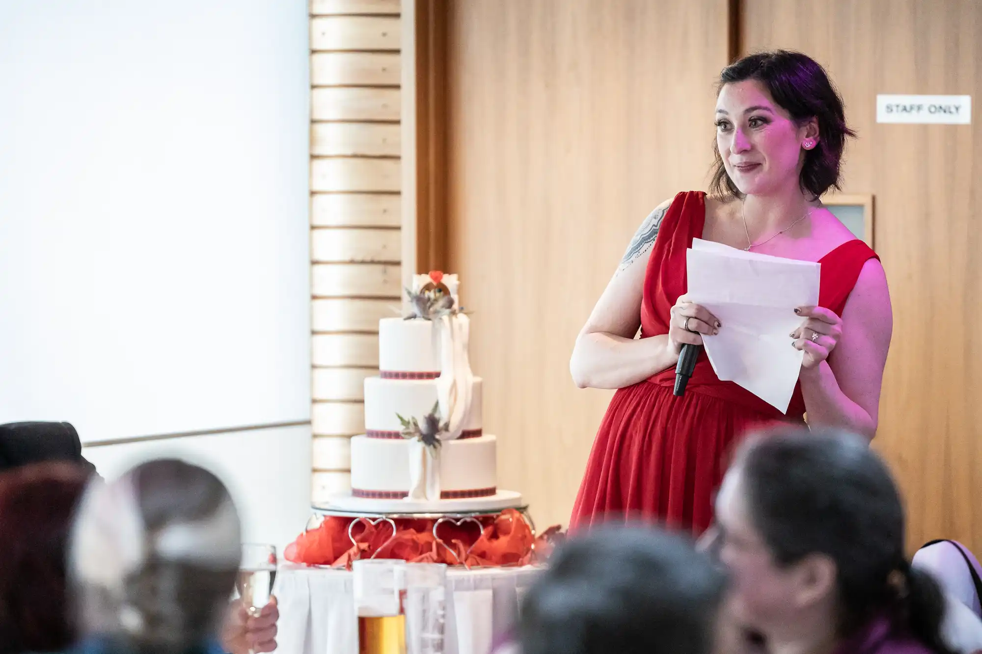 A woman in a red dress holds a microphone and papers, smiling as she stands near a three-tier white wedding cake decorated with flowers. She faces seated guests in a bright room, with a “Staff Only” sign visible on a wooden door behind her.