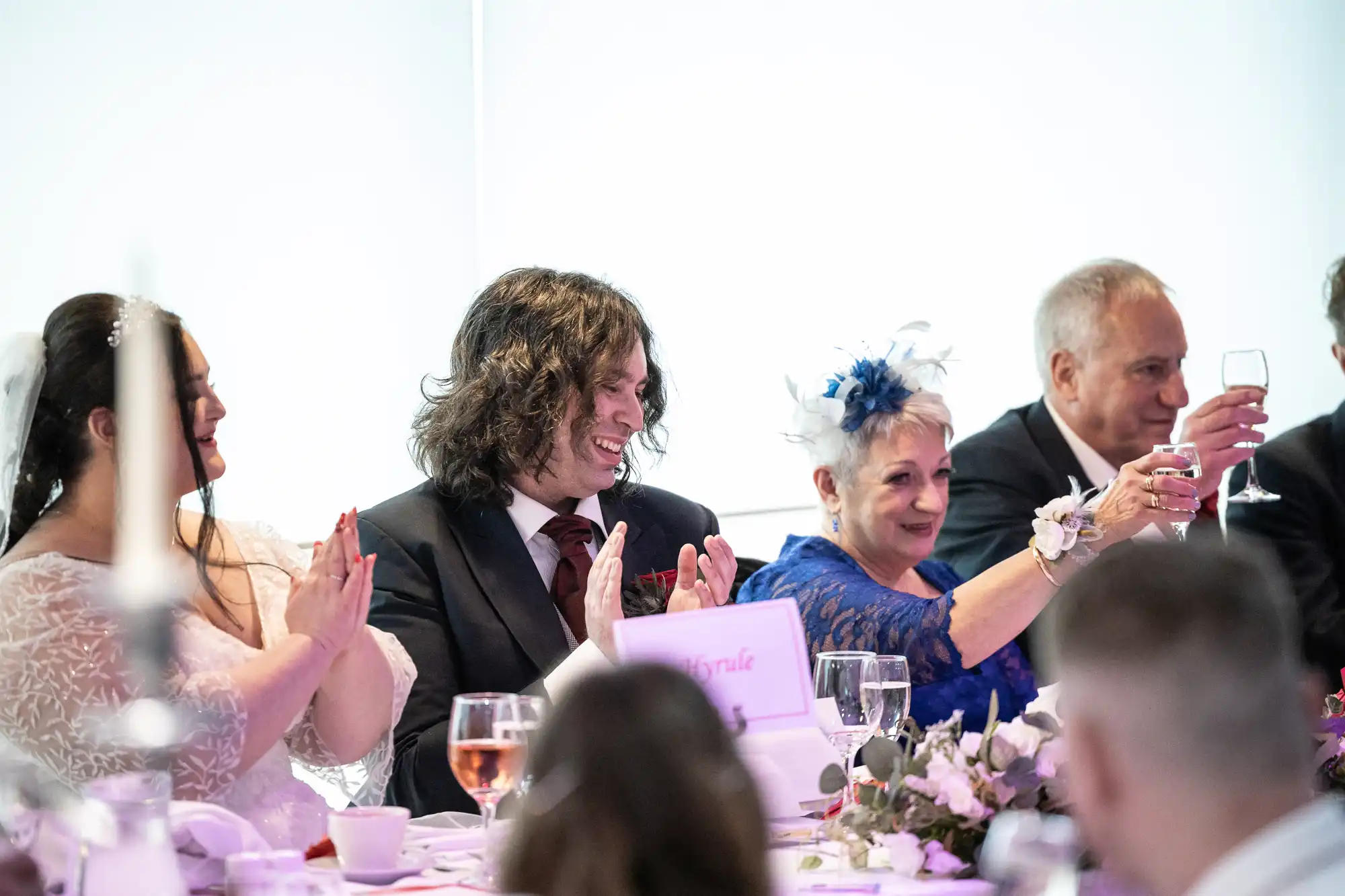 A wedding reception scene shows guests at a table clapping and smiling. A woman in white sits beside a man in a suit with long curly hair. Next to him, a woman in blue with a fascinator raises her glass in a toast. The table is decorated with flowers and drinks.