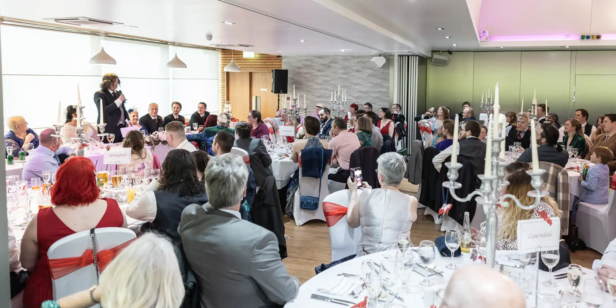 A large group of people dressed in formal attire are seated at round tables in a banqueting hall, attentively listening to a person speaking at the front of the room. The tables are decorated with white cloths, glasses, and tall candlesticks.