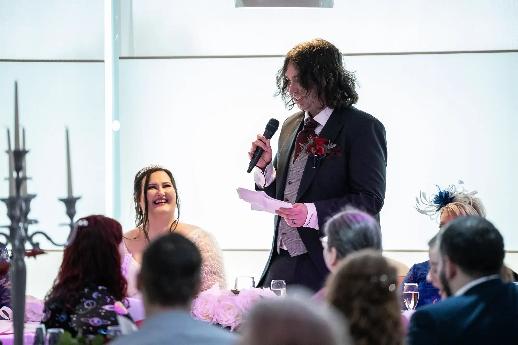 A man in a suit with long dark hair gives a speech with a microphone at a formal event. A woman in a white dress, possibly a bride, sits beside him smiling. Several guests are seated around them, watching and listening attentively.