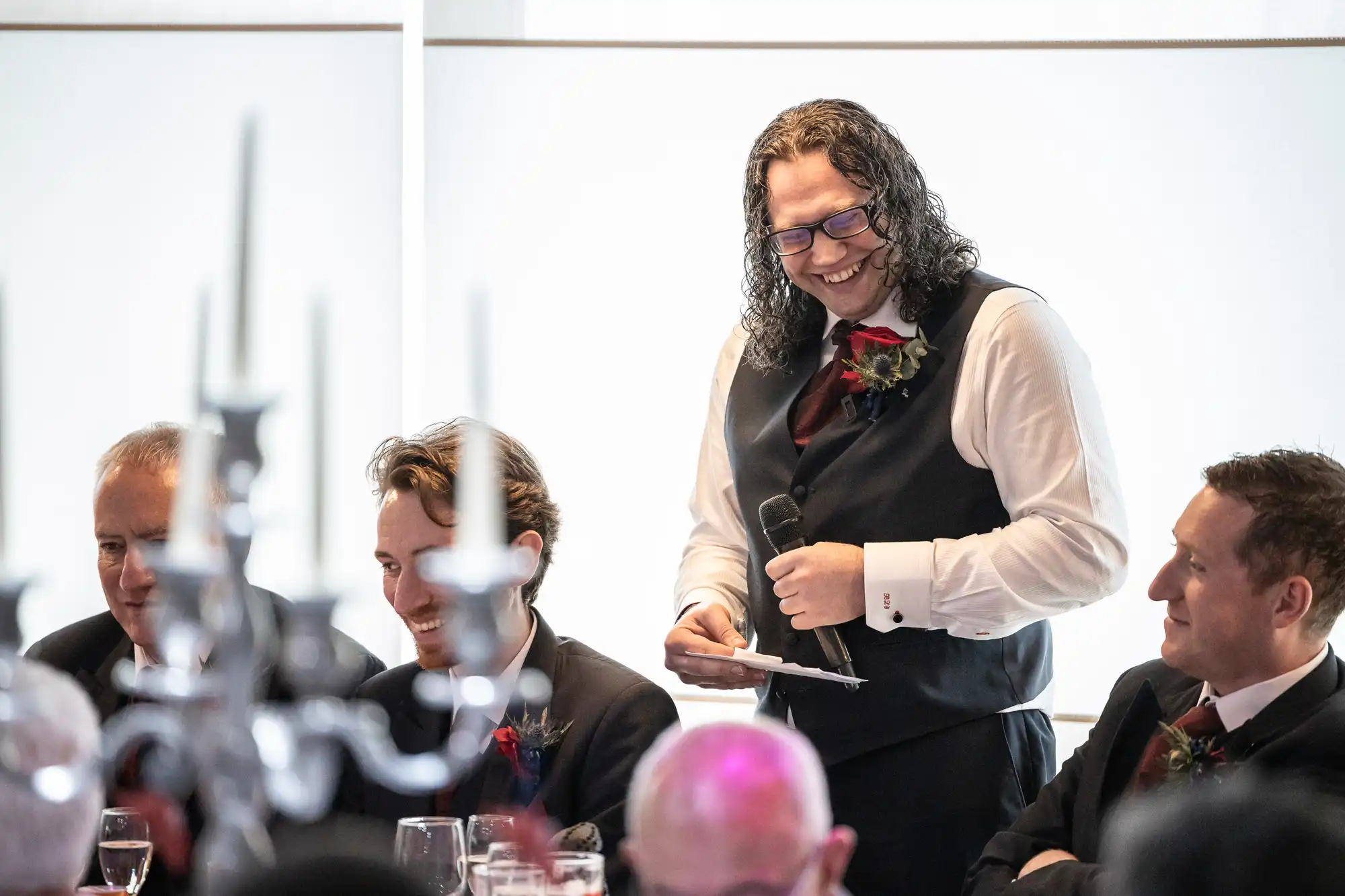 A man with long curly hair, glasses, and a black waistcoat stands smiling with a microphone and notes. He is speaking to seated men in suits who are also smiling or laughing at a formal event, possibly a wedding reception, with a candelabrum in the foreground.