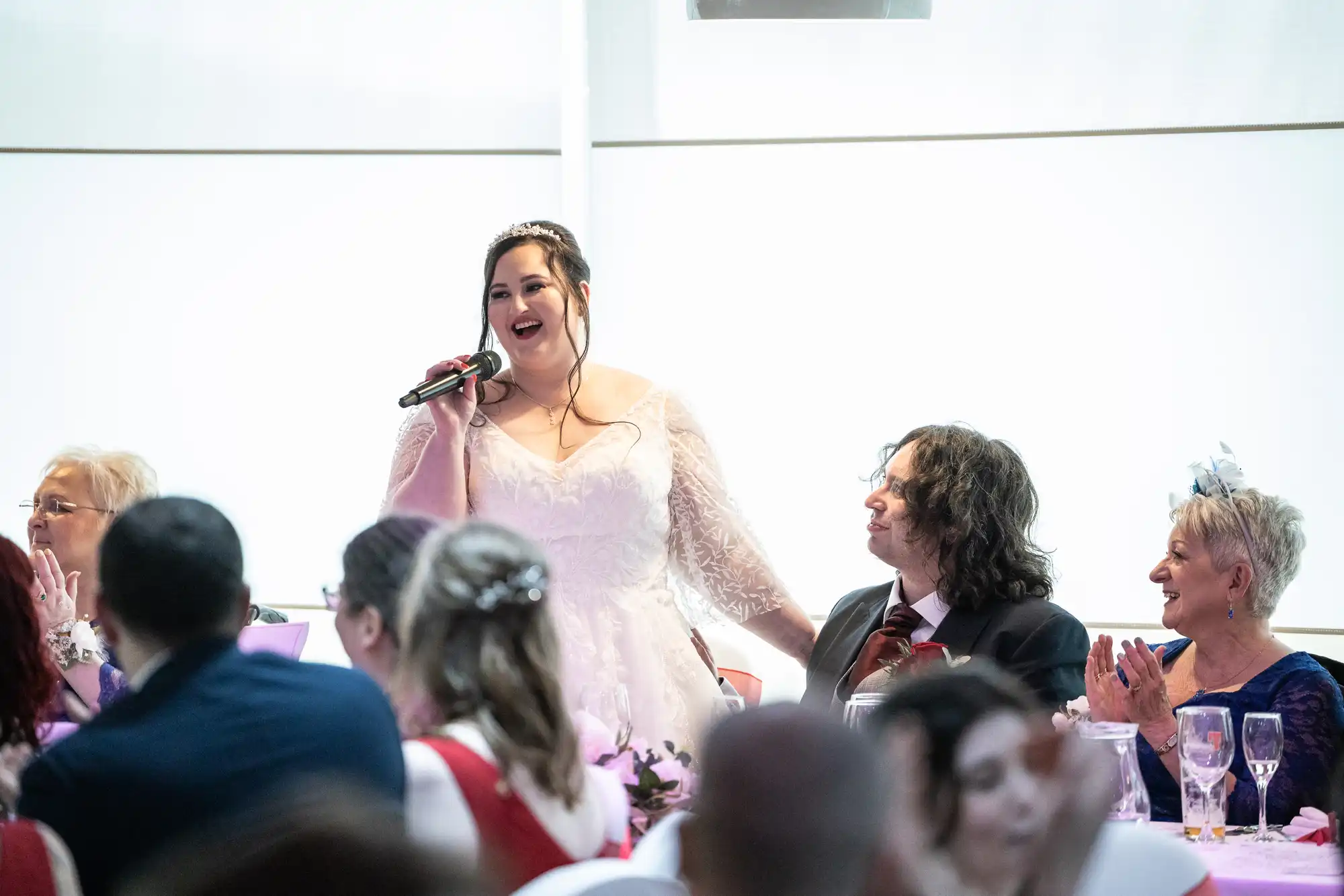 A bride in a white lace dress stands smiling with a microphone, giving a speech at a wedding reception. Guests, seated around decorated tables, smile and applaud. The groom sits to her left, looking up at her. The scene is bright and joyful.
