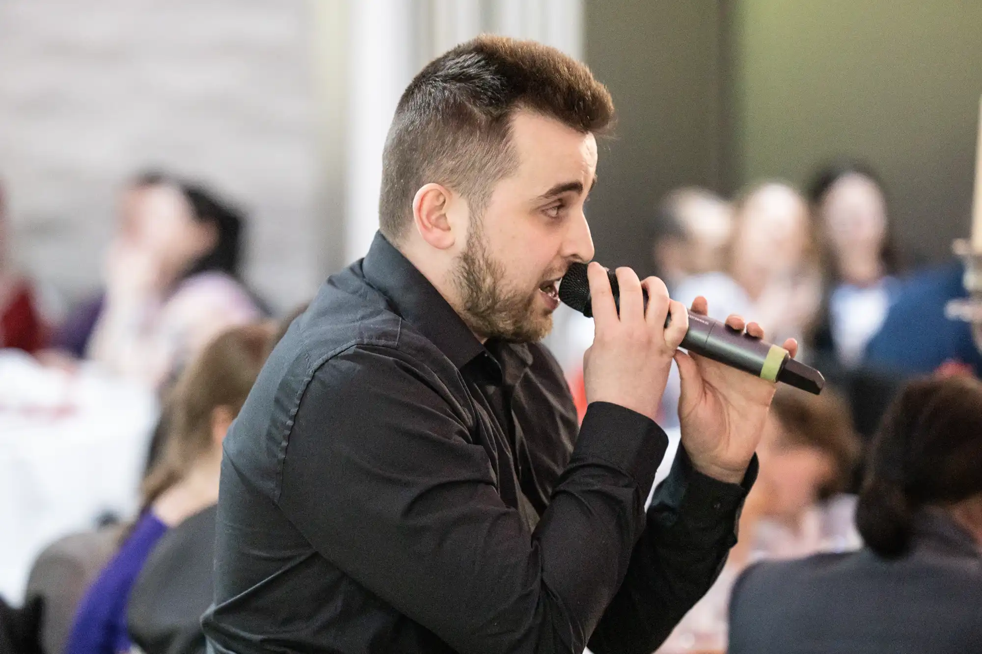 A man with short dark hair and a beard, wearing a black shirt, holds a microphone while speaking or singing. He is indoors, with people seated and blurred in the background, suggesting a social or formal event.