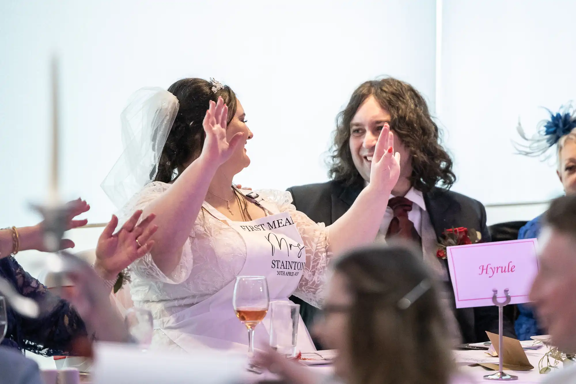 A bride in a white dress and veil raises her arms, smiling joyfully at a wedding reception. A man in a suit with long hair sits beside her, smiling. Guests surround them, and a table sign reading “Hyrule” is visible in front. Warm, celebratory atmosphere.