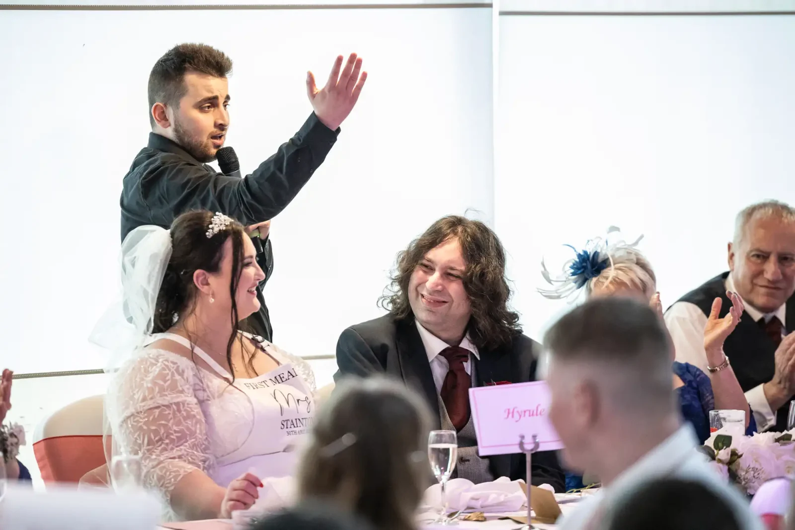 A singing waiter in a black shirt stands and speaks with a microphone, gesturing with his hand. Seated at a decorated table, a bride in white smiles at him, and guests, including a man in a suit, listen and react, suggesting a wedding reception speech.