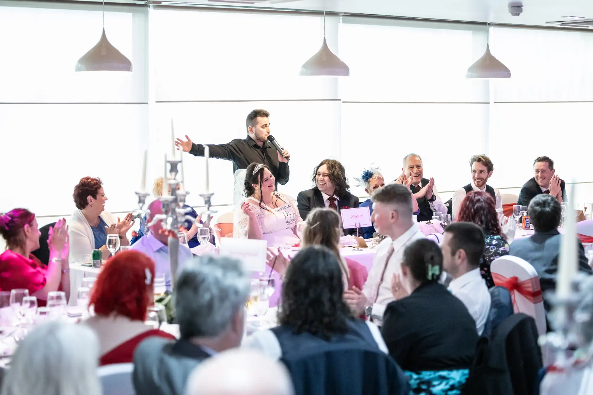 A lively wedding reception scene shows a man standing with a microphone, addressing seated guests at a long banquet table decorated with pink and white accents. Guests are smiling, applauding, and dressed in formal attire. Large windows and hanging lights brighten the room.