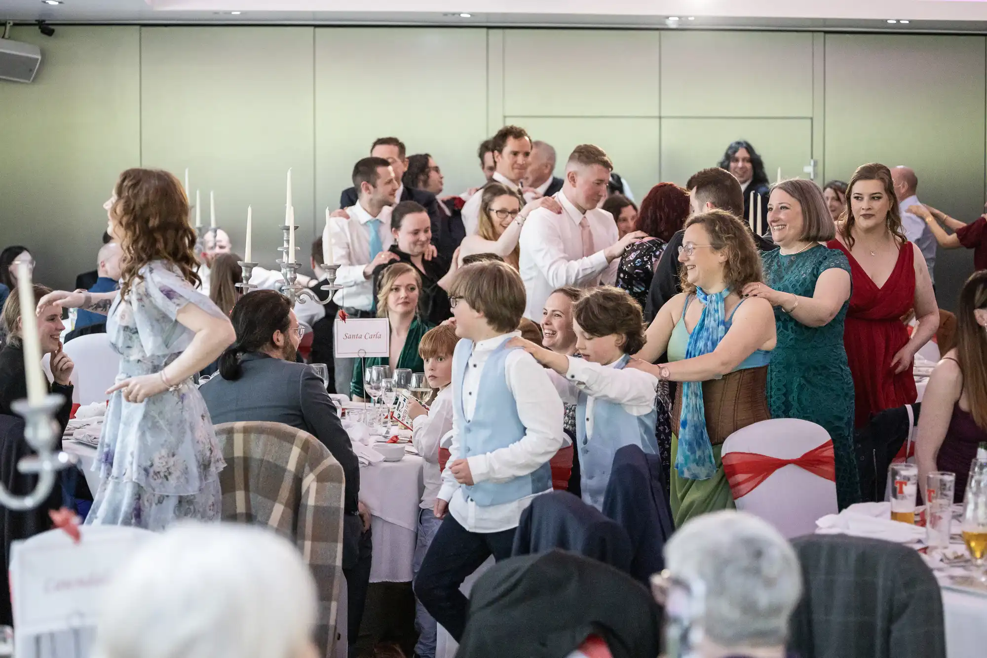 A lively group of people at a formal event form a conga line around banquet tables, smiling and laughing. Guests wear semi-formal attire, and tables are set with white cloths, glasses, and candles. The atmosphere is festive and joyful.