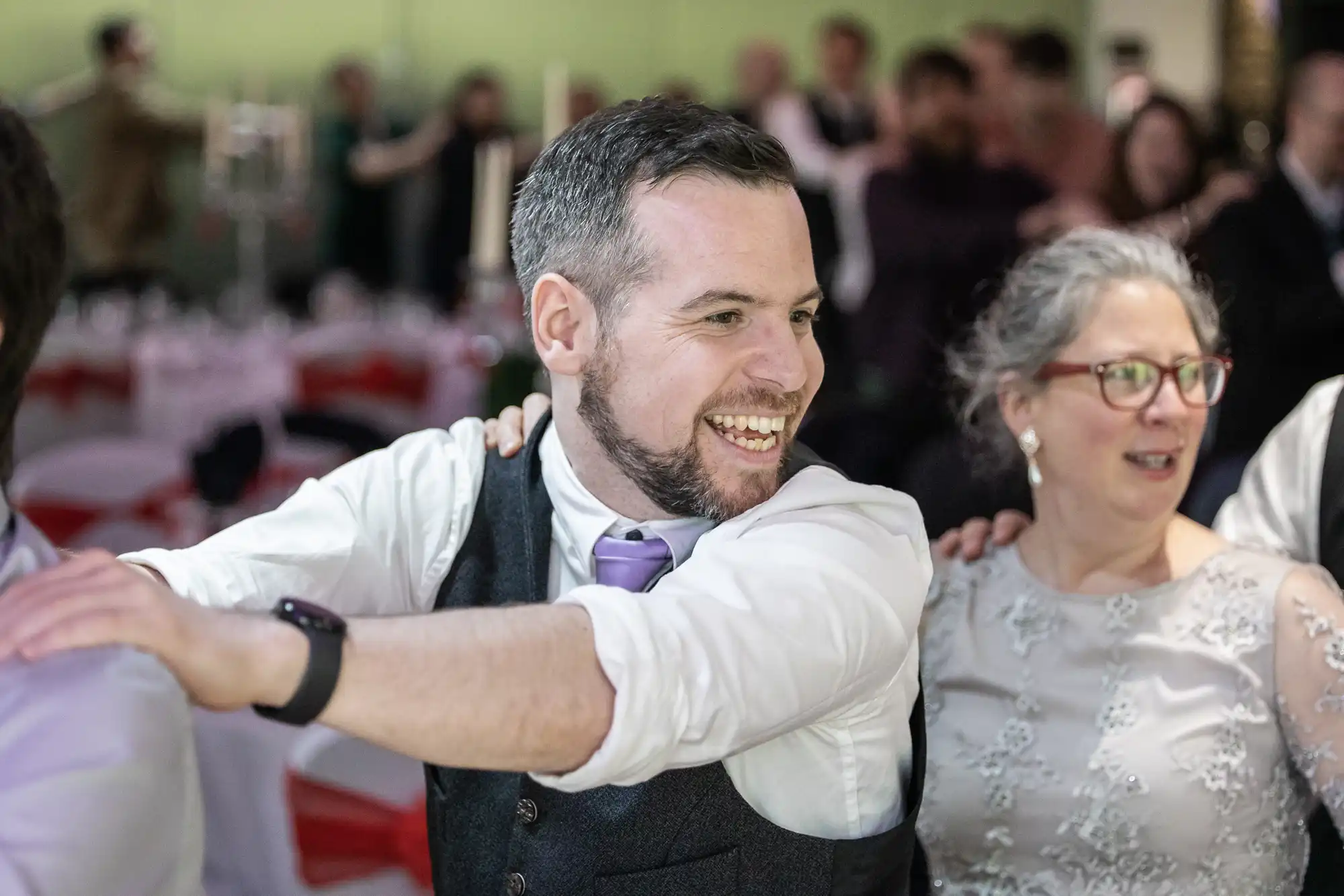 A smiling man in a waistcoat and lavender tie stands in a festive indoor setting, with his hands on the shoulders of a woman in a grey lace dress beside him. They’re part of a conga line, with blurred people and decorated tables in the background.