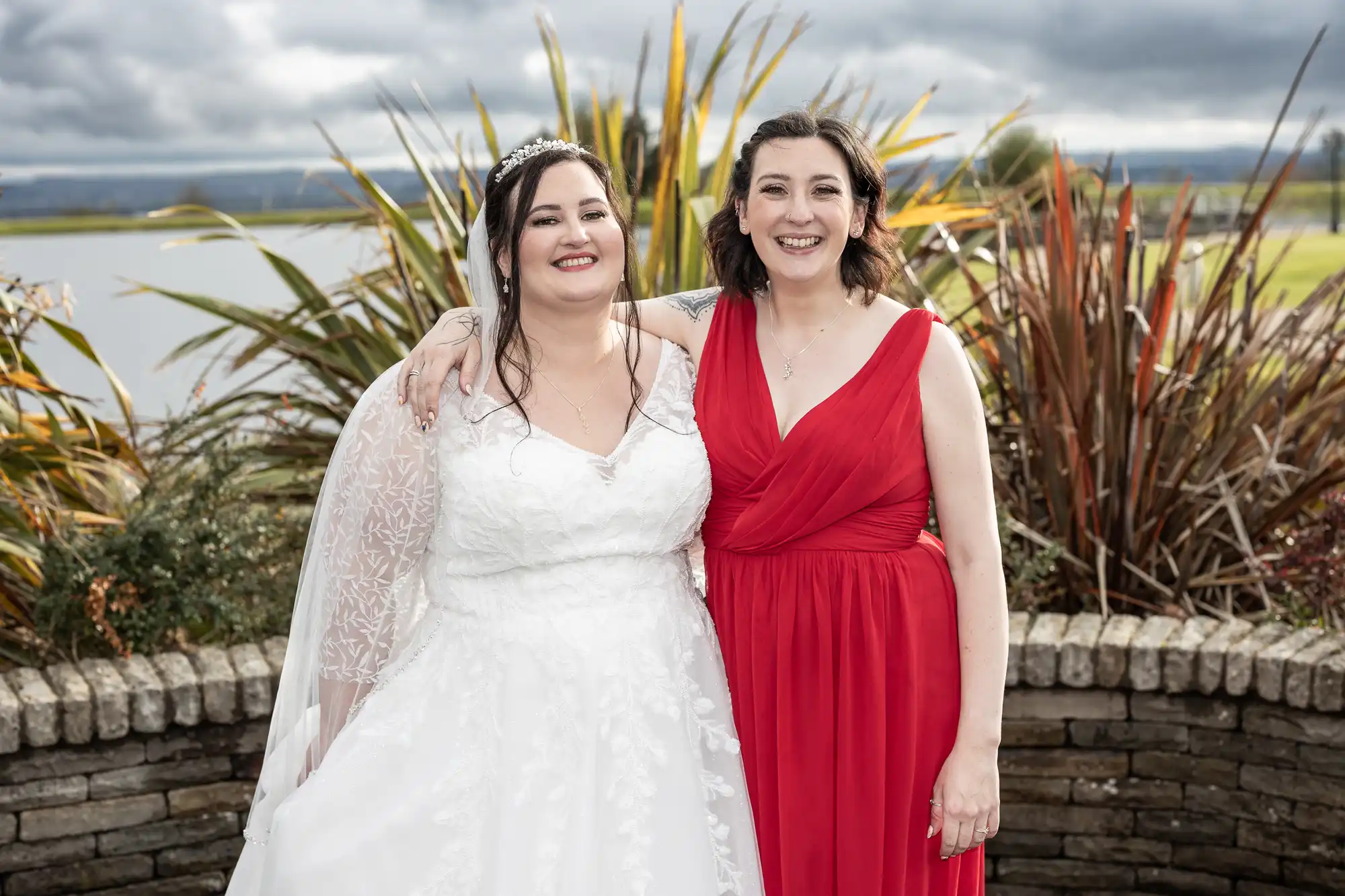 Two women stand outdoors, smiling. The woman on the left wears a white wedding dress and veil, while the woman on the right wears a sleeveless red dress. They have their arms round each other. Behind them are tall plants, a stone wall, and a lake under a cloudy sky.