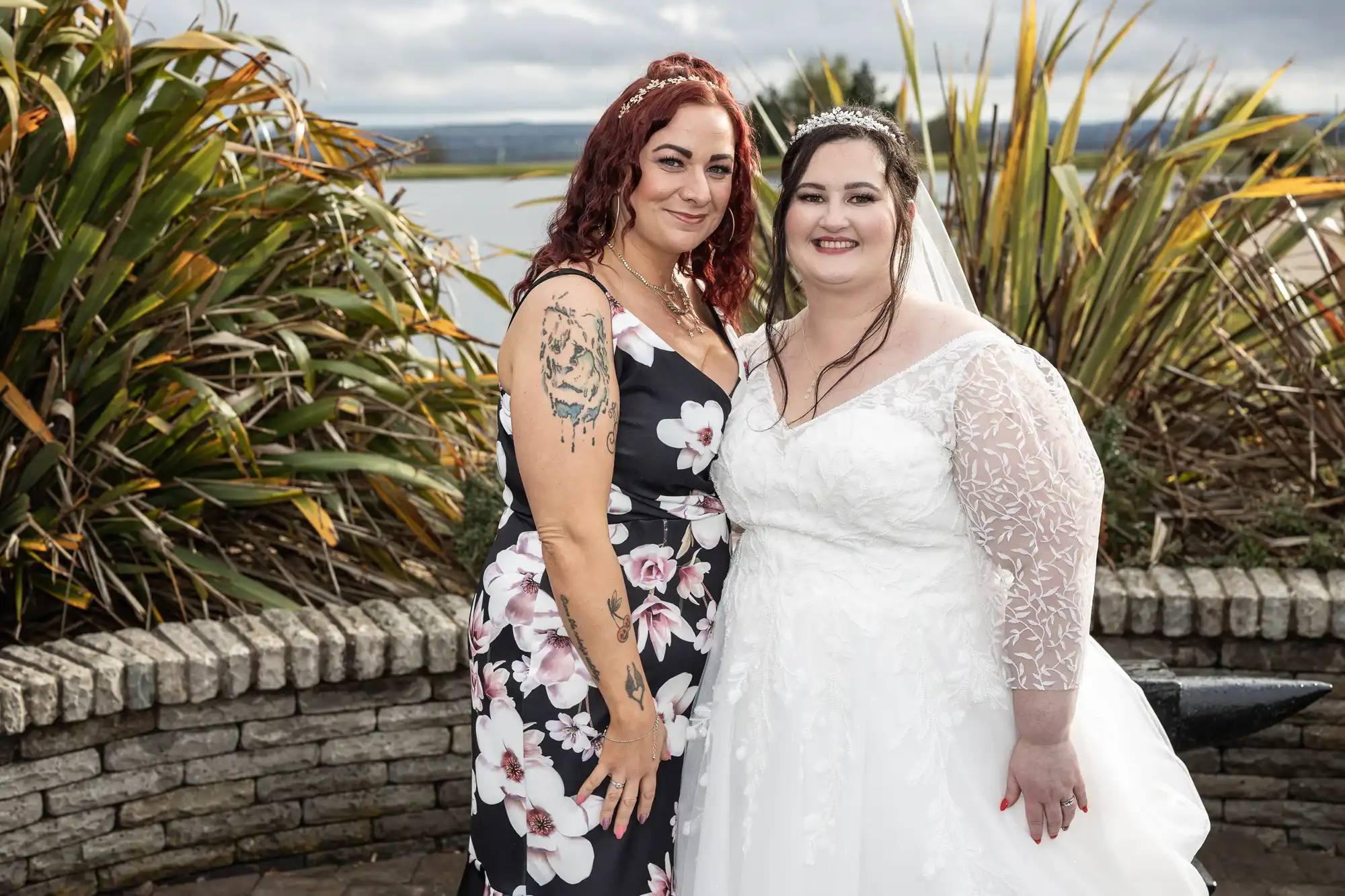 Two women pose together outdoors. One wears a white wedding dress with lace sleeves and a tiara; the other wears a black floral dress, showing tattooed arms. They smile, with greenery, stone wall, and water in the background under a cloudy sky.