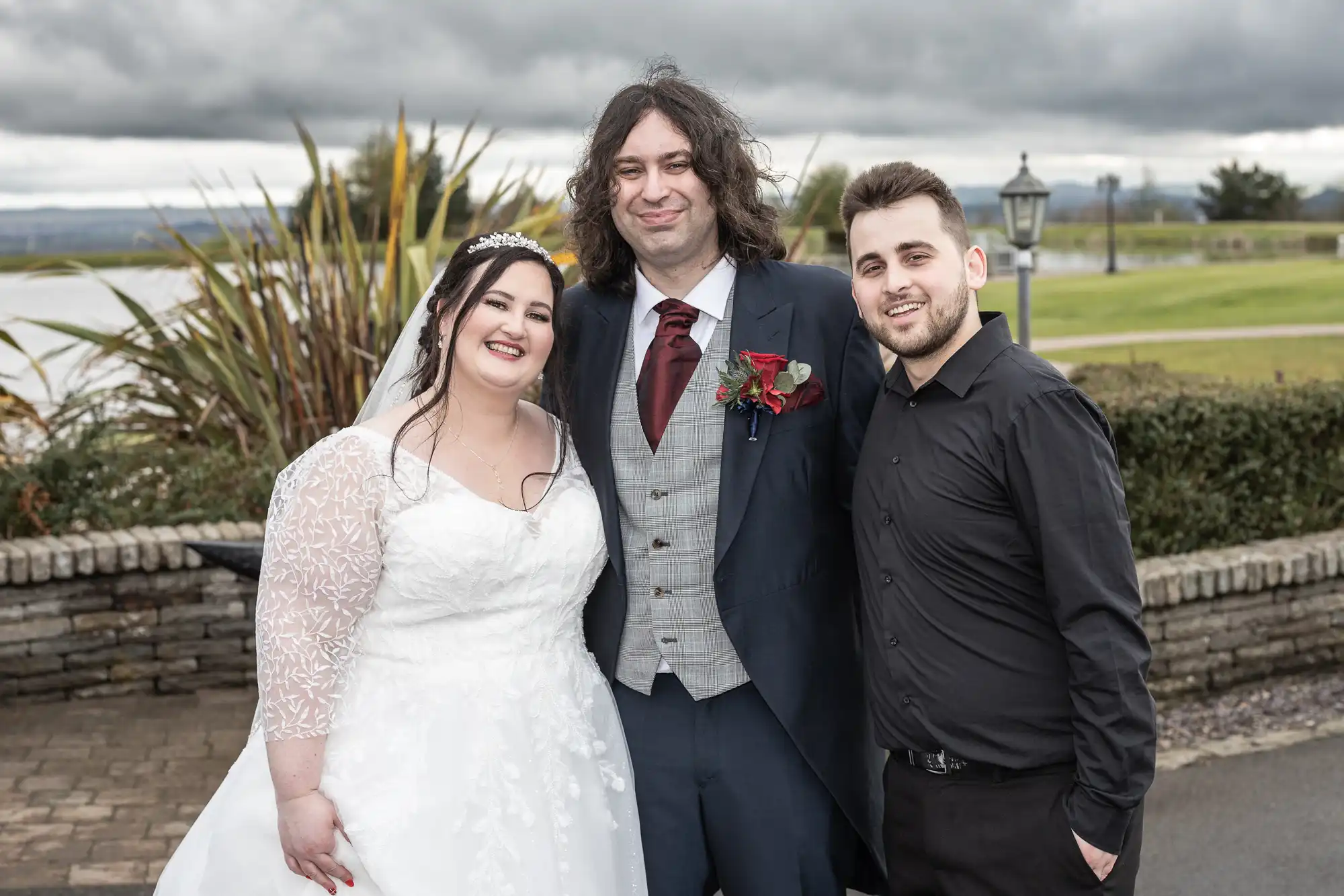 A bride in a white dress, a groom in a grey suit with a red tie, and a man in a black shirt pose smiling outdoors. Behind them are green lawns, a stone wall, plants, and a cloudy sky.