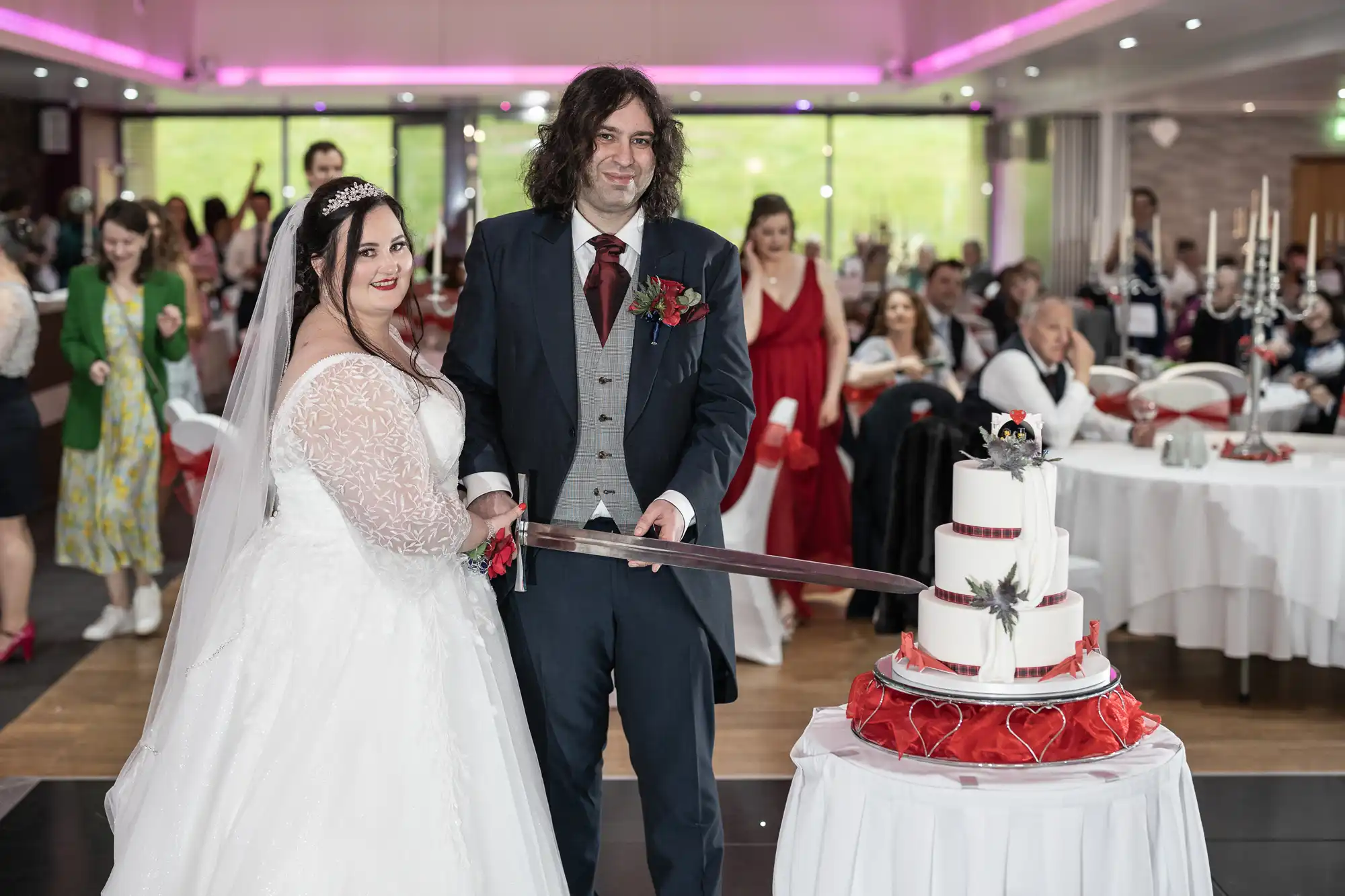 A bride and groom in formal wedding attire smile as they cut a white three-tiered cake with a large sword. The cake is decorated with red ribbons and feathers. Guests watch from round tables in a spacious, modern venue with pink lighting.