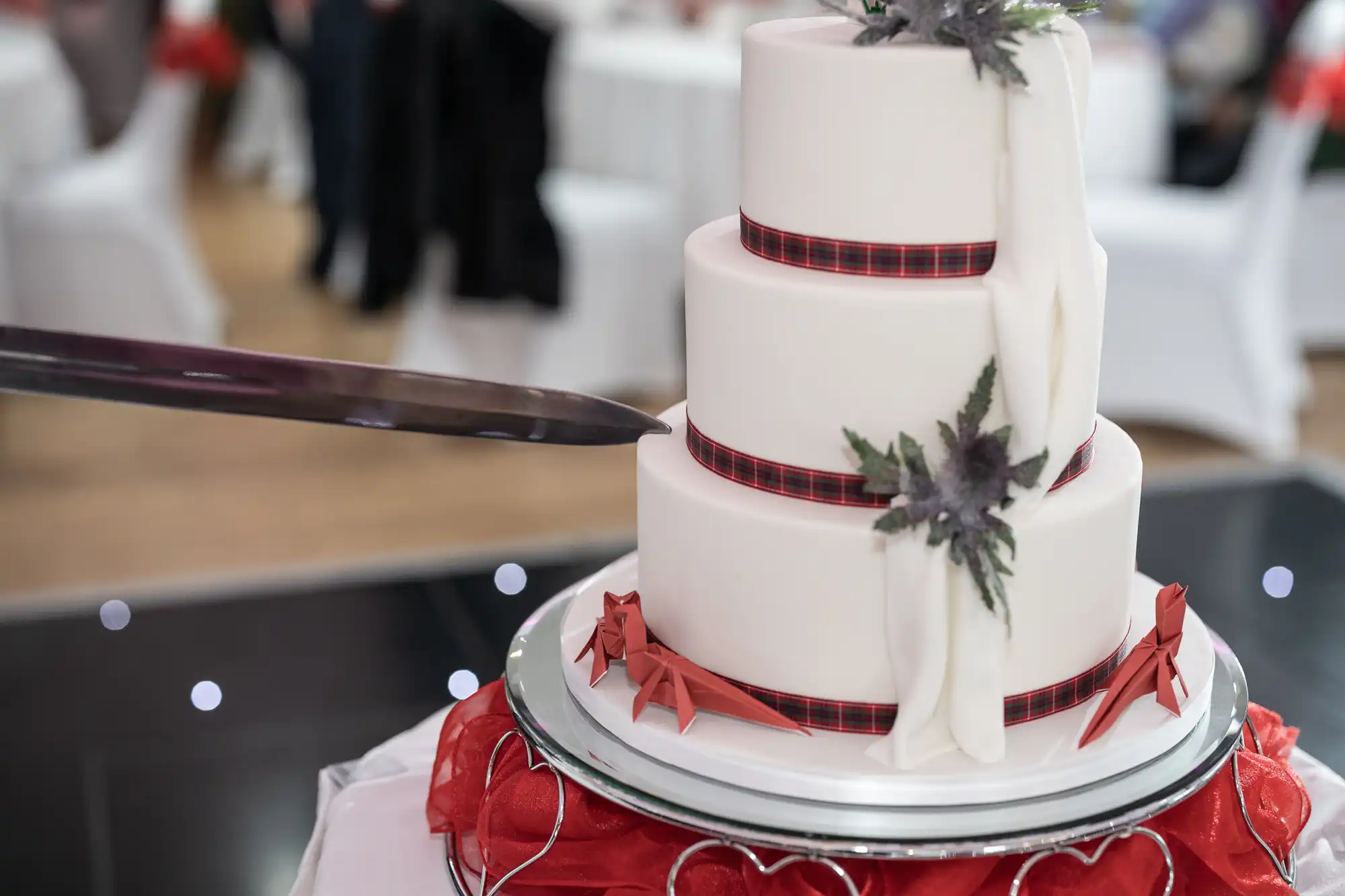A close-up of a three-tier white wedding cake with red and black ribbon accents, white drapery details, and floral decorations. A large knife is positioned to cut the cake, which sits on a reflective silver stand with red fabric and white heart shapes.