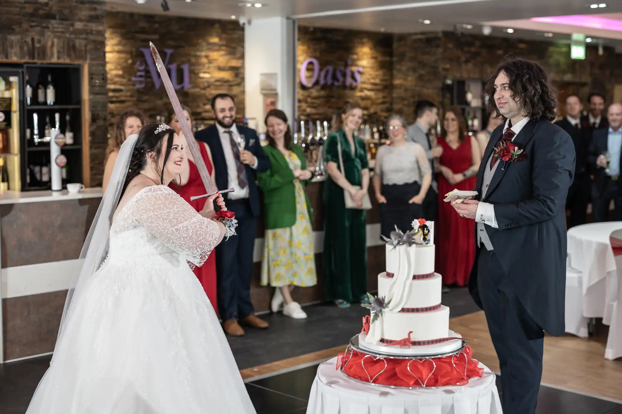 A bride in a white dress playfully points a cake knife at the groom, who smiles holding a plate, as they stand by a three-tier wedding cake. Guests in colourful outfits watch and smile in a warmly lit reception hall.