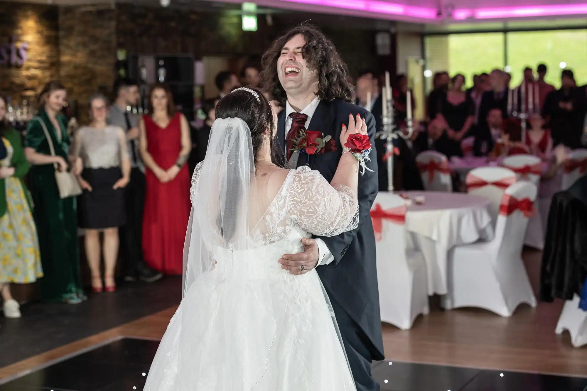 A bride and groom share a joyful first dance at their wedding reception. The groom laughs with his mouth open, while the bride faces him. Guests in formal attire watch them, and tables with white cloths and red bows surround the dance floor.