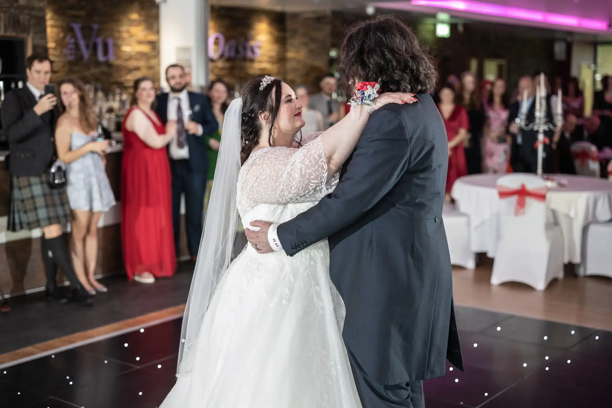 A bride in a white dress and veil dances with a groom in a dark suit on a black dance floor, smiling at each other. Guests in formal wear watch and take photos in the background. The venue has purple lighting and decorated tables.