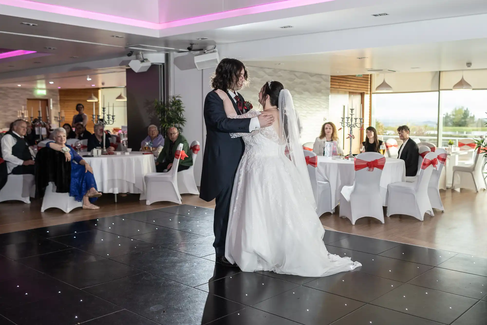 A bride and groom share their first dance on a black-tiled floor in a brightly lit reception hall. Guests sit at round tables with white cloths and red sashes, watching the couple. The bride wears a white gown and veil; the groom wears a dark suit with long hair.