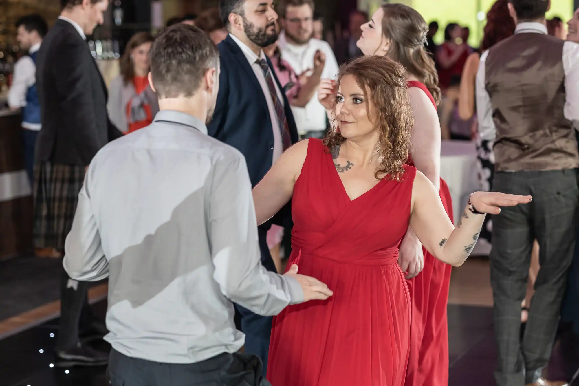 A woman in a red dress dances and gestures with her hands while facing a man in a light grey shirt. Other people in formal attire socialise and dance in the background at what appears to be a lively indoor event or celebration.
