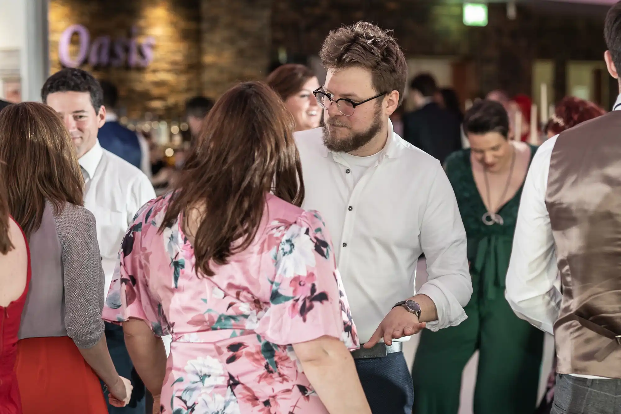 A bearded man in glasses and a white shirt dances with a woman in a pink floral dress at a lively indoor event. Other guests, some smiling and dressed formally, mingle and dance in the softly lit background near a sign that reads “Oasis”.