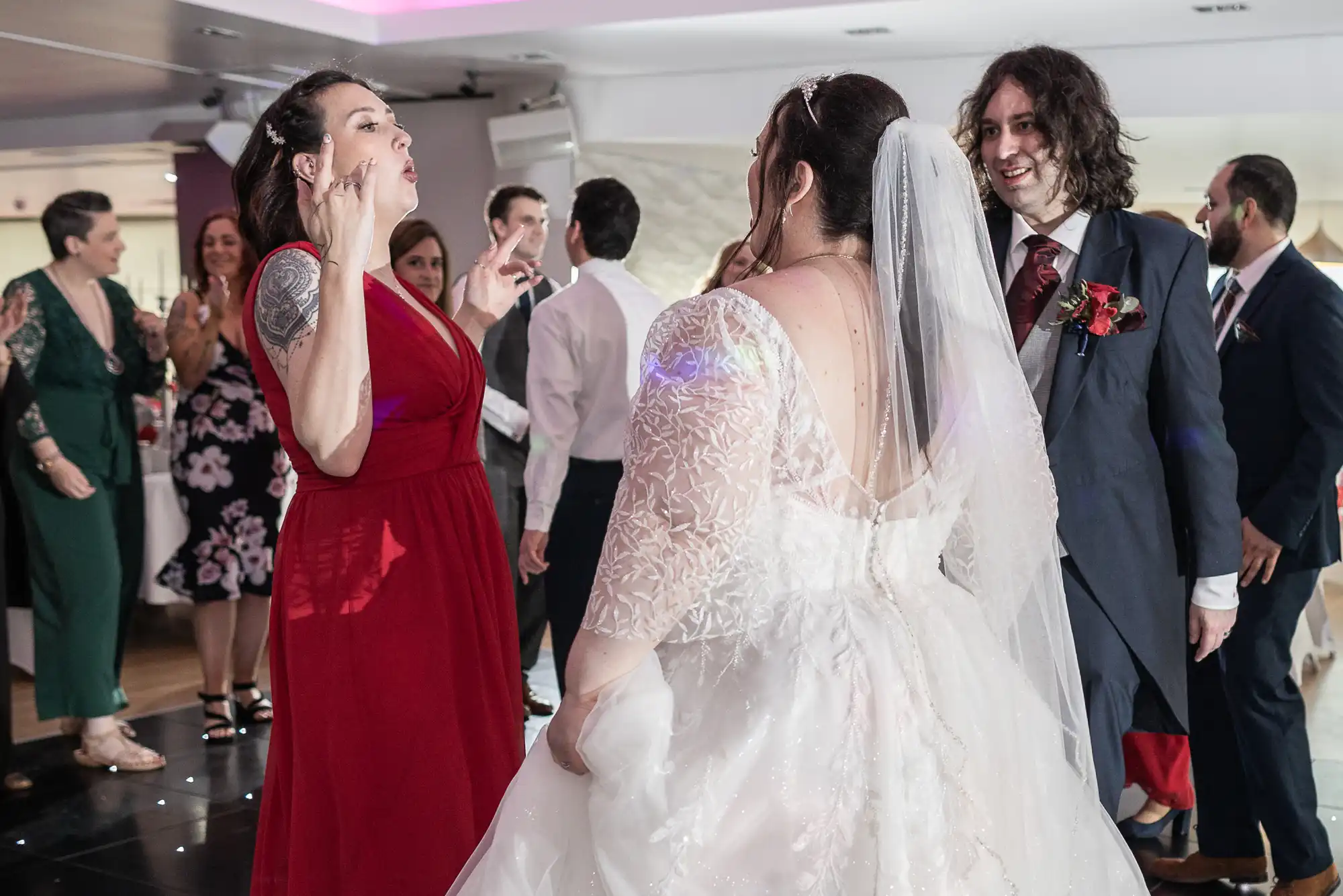 A bride in a white dress and veil dances with guests at a wedding reception. A woman in a red dress gestures animatedly, whilst a man in a blue suit with long hair smiles. Other guests in formal attire socialise in the background under soft lighting.