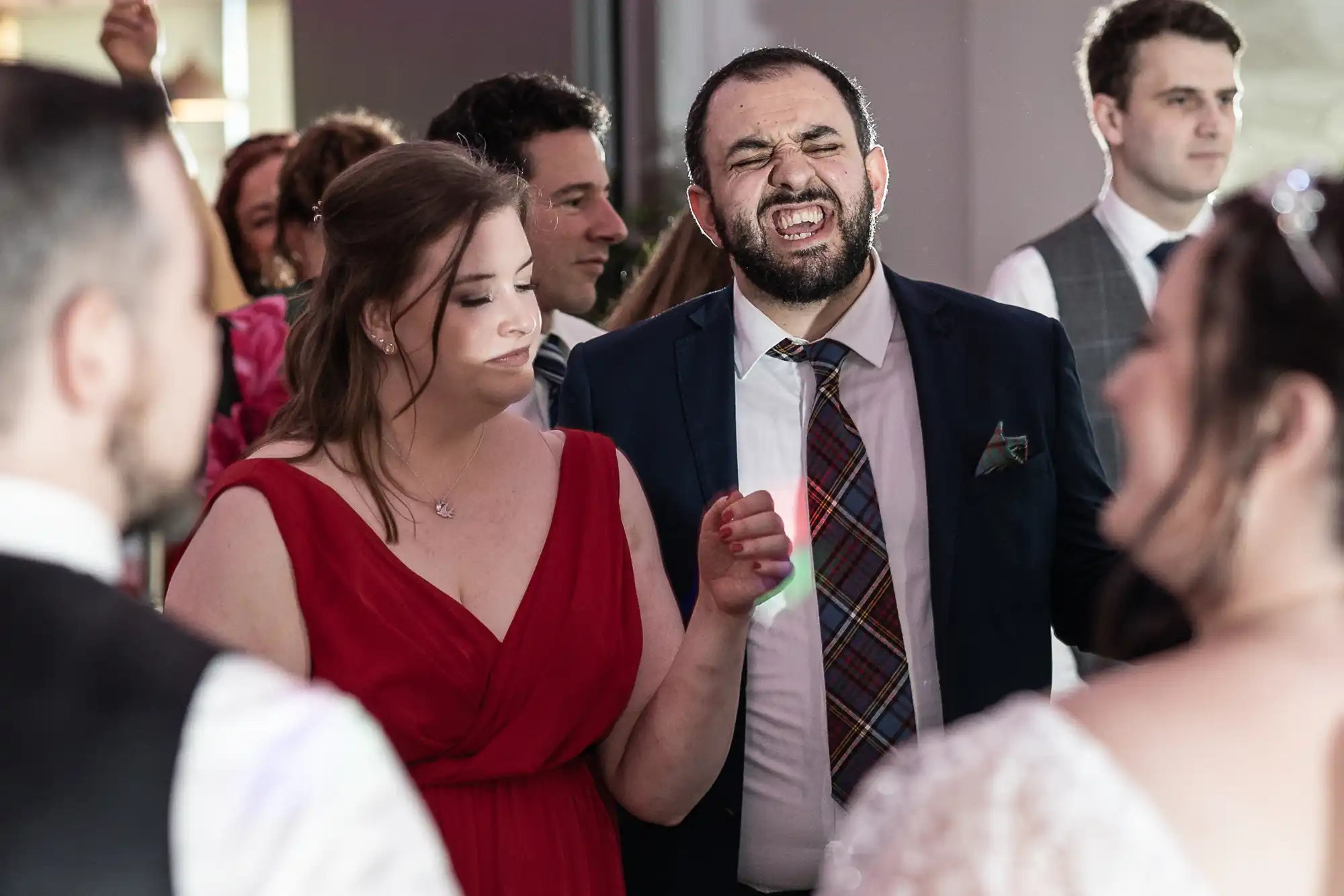 A man in a suit and checked tie sings passionately whilst holding hands with a woman in a red dress at a lively indoor event. Other guests, some smiling, are visible in the background, all dressed formally, suggesting a celebration or wedding.