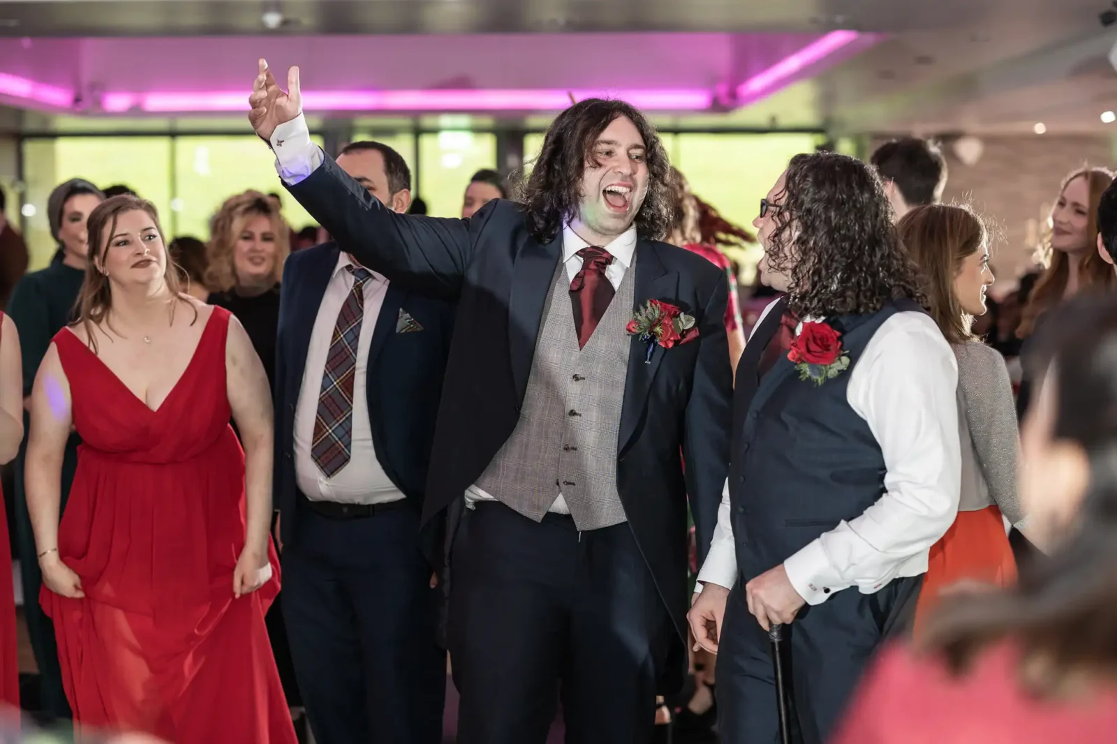 Two men in suits, one with a red buttonhole and another with a red rose, stand joyfully among a crowd at an indoor event. The man in the centre raises his arm, smiling widely. Guests in formal attire and red dresses surround them under pink lighting.