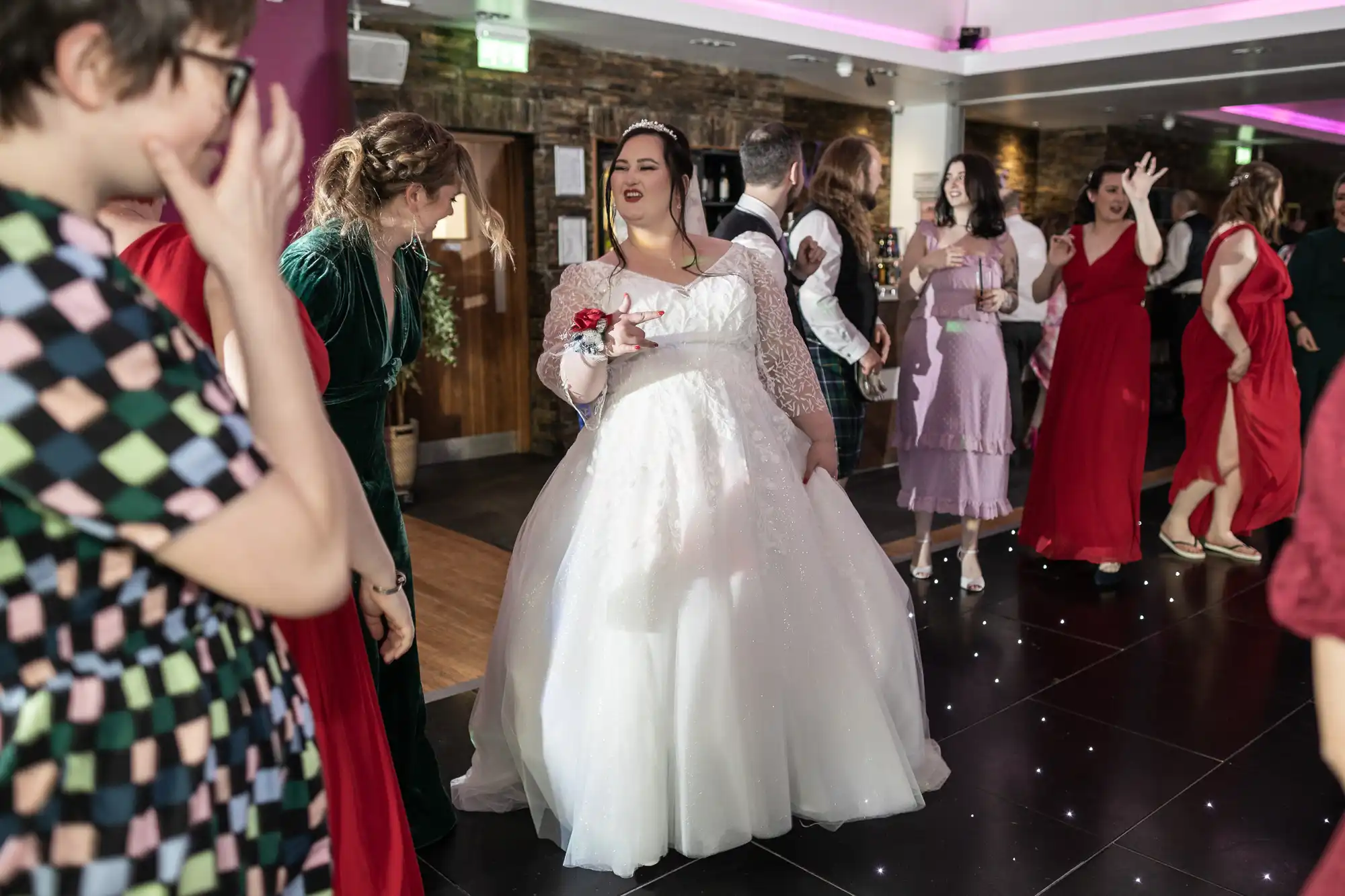 A bride in a white dress and veil smiles and gestures while dancing with guests at a lively indoor wedding reception. People in colourful dresses chat, laugh, and dance on a shiny black floor under festive lighting. The atmosphere is joyful and celebratory.