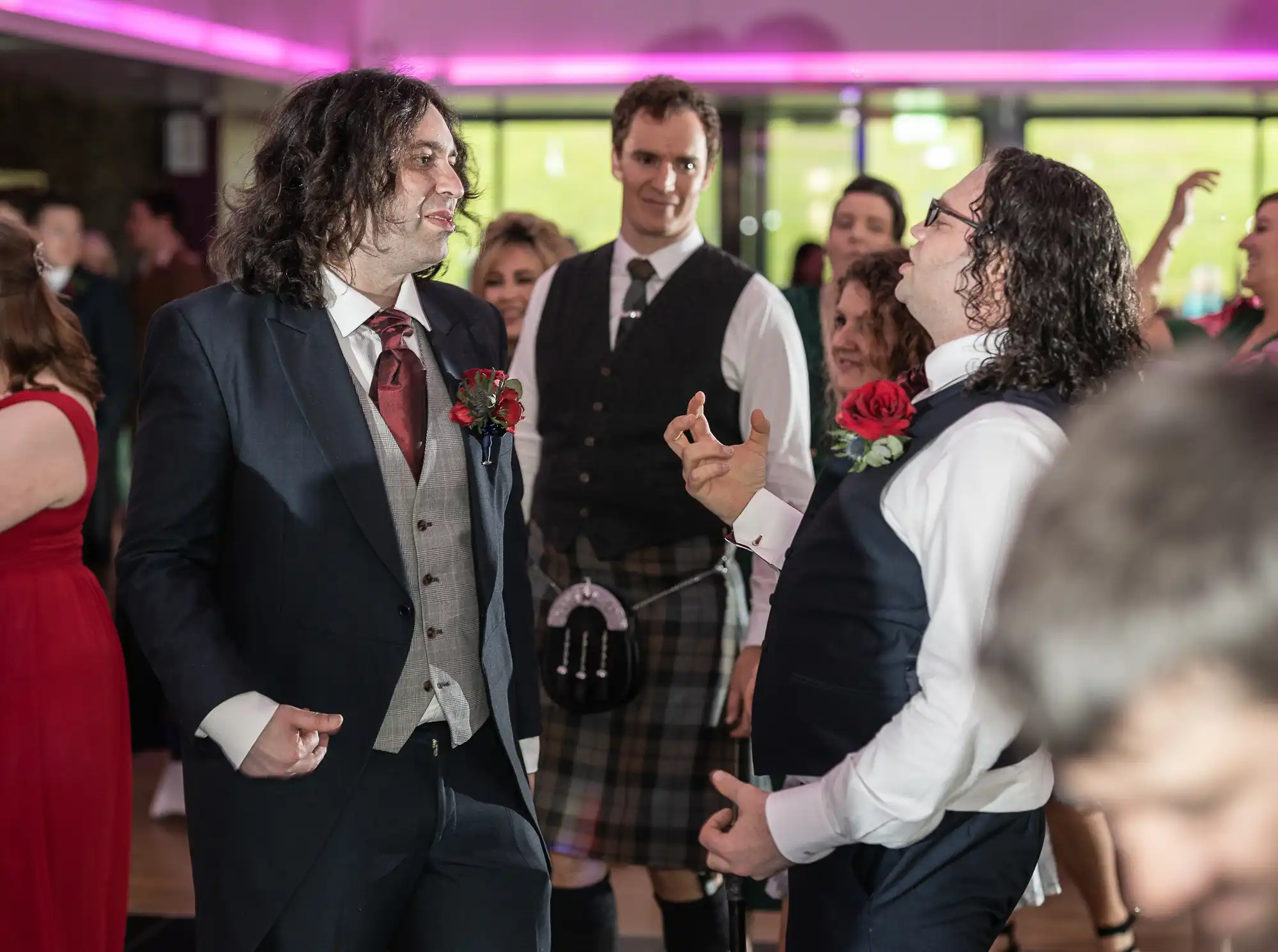 Several people are at a wedding reception. Two men in suits are dancing and smiling, while a man in a kilt and waistcoat stands between them. In the background, guests in formal attire watch and enjoy the lively, joyful atmosphere. Pink lighting adds a festive touch.