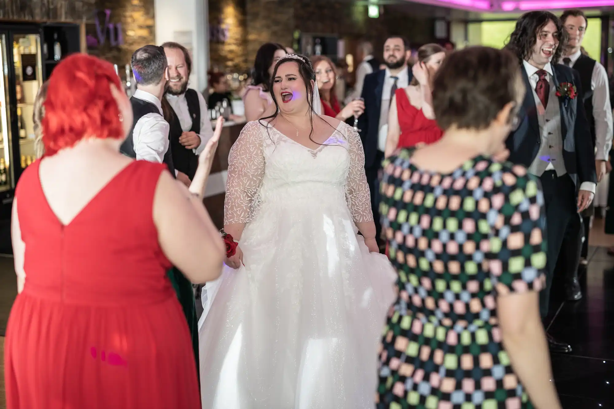 A joyful bride in a white gown with lace sleeves smiles brightly whilst surrounded by wedding guests dancing. People wear colourful dresses and suits, creating a lively, celebratory atmosphere inside a warmly lit reception hall.