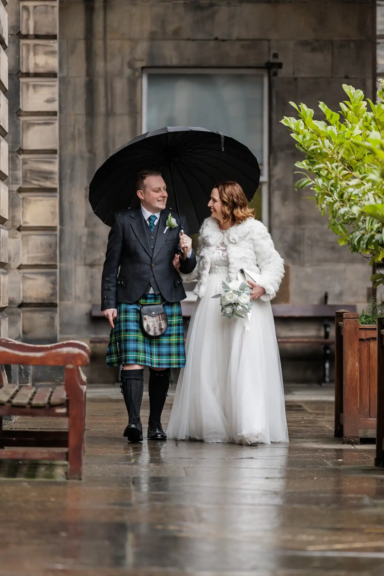 A smiling groom in a Scottish kilt and a bride in a white dress and fur stole walk arm in arm under a black umbrella on a wet stone path, with greenery and stone buildings in the background.