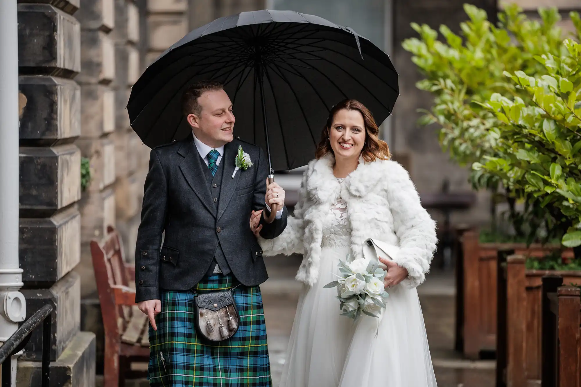 A smiling bride in a white dress and faux fur jacket walks arm-in-arm with a groom in a kilt and jacket. He holds a black umbrella over them as they walk outdoors on a rainy day, surrounded by stone buildings and greenery.