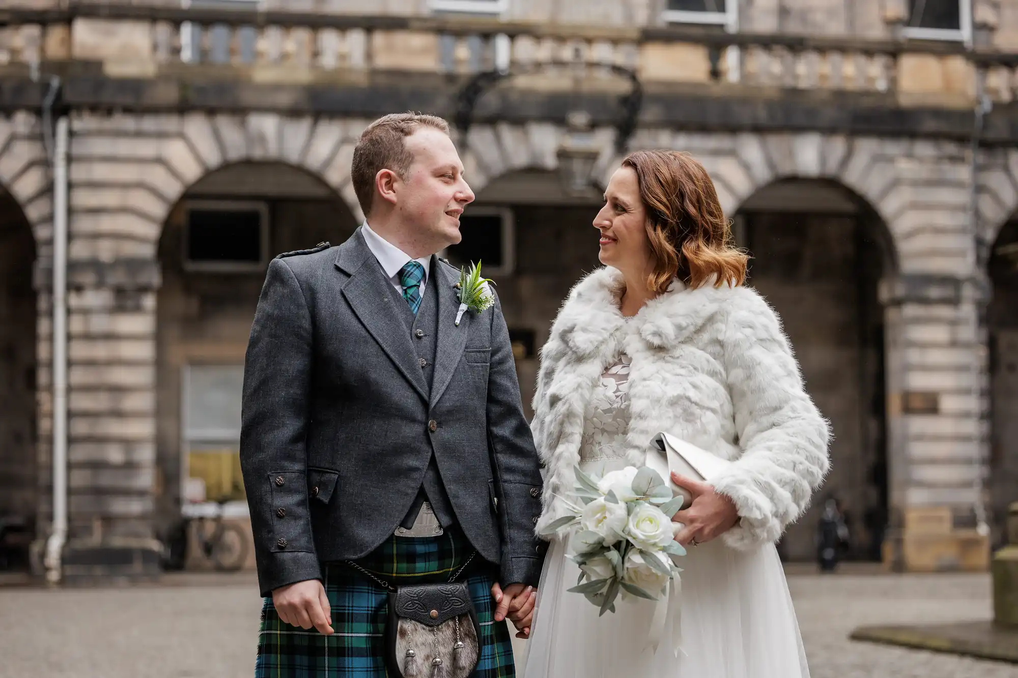 A smiling couple stands outdoors in front of a stone building. The man wears a traditional Scottish kilt outfit with a sporran. The woman wears a white dress with a fur jacket and holds a bouquet of white flowers. They are holding hands and looking at each other.