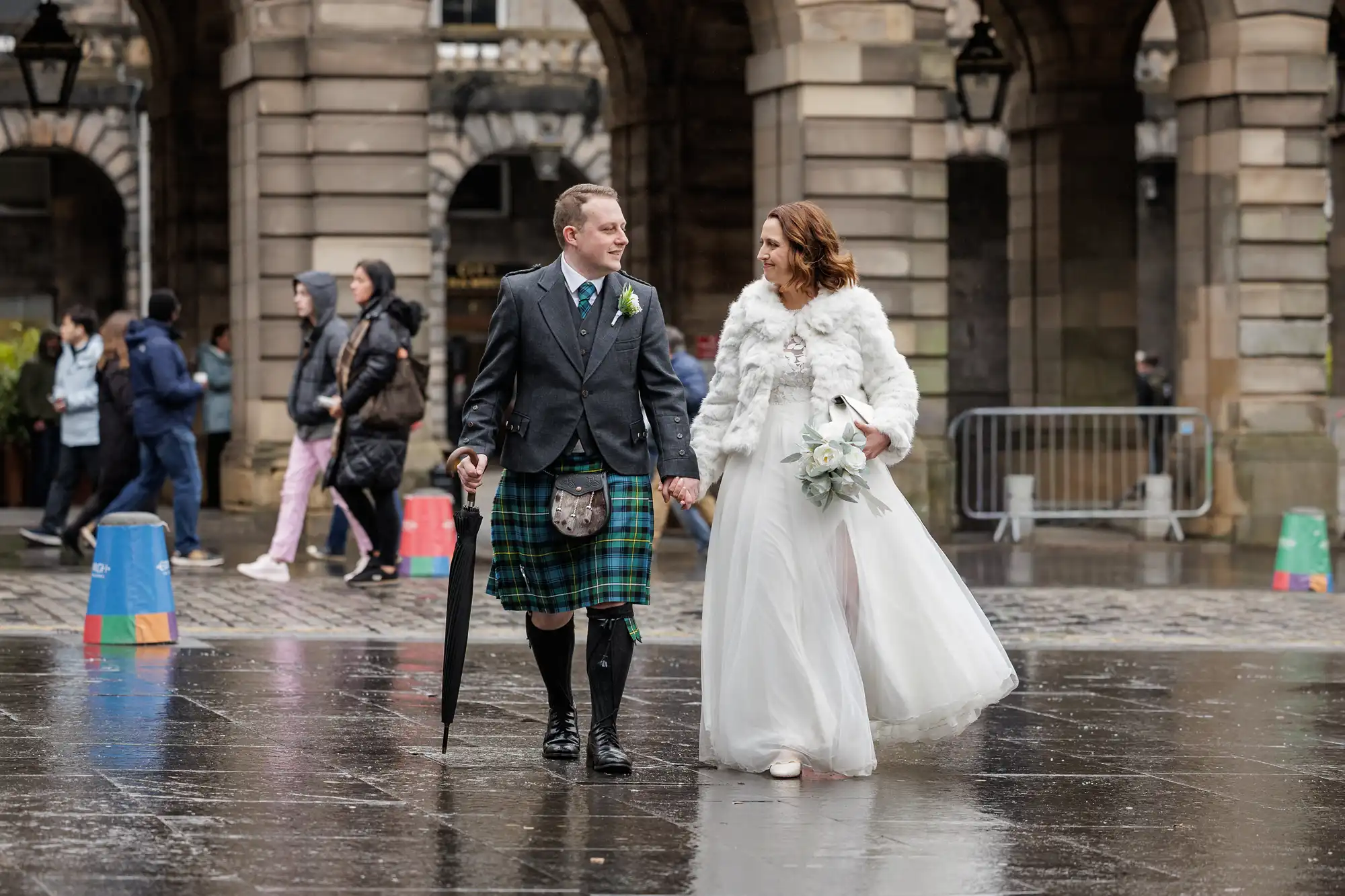 A bride in a white dress and fur stole holds a bouquet and walks hand-in-hand with a groom in a dark jacket and green tartan kilt. They’re on a wet stone street, looking at each other, with people and stone arches blurred in the background.