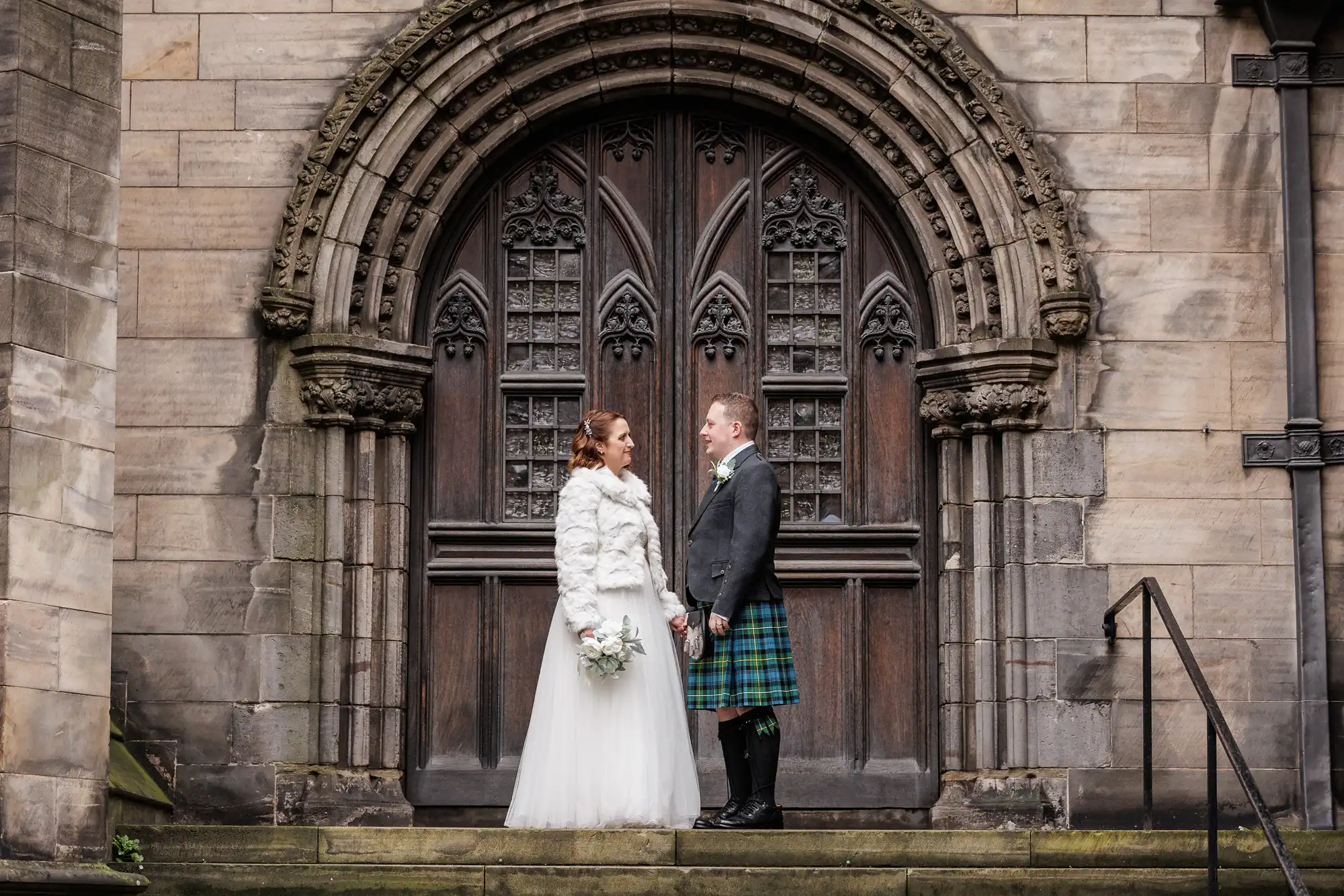 A bride in a white gown and jacket holds a bouquet, standing beside a groom in a tartan kilt and jacket. They face each other on stone steps in front of an ornate arched wooden church door set in a gothic stone façade.