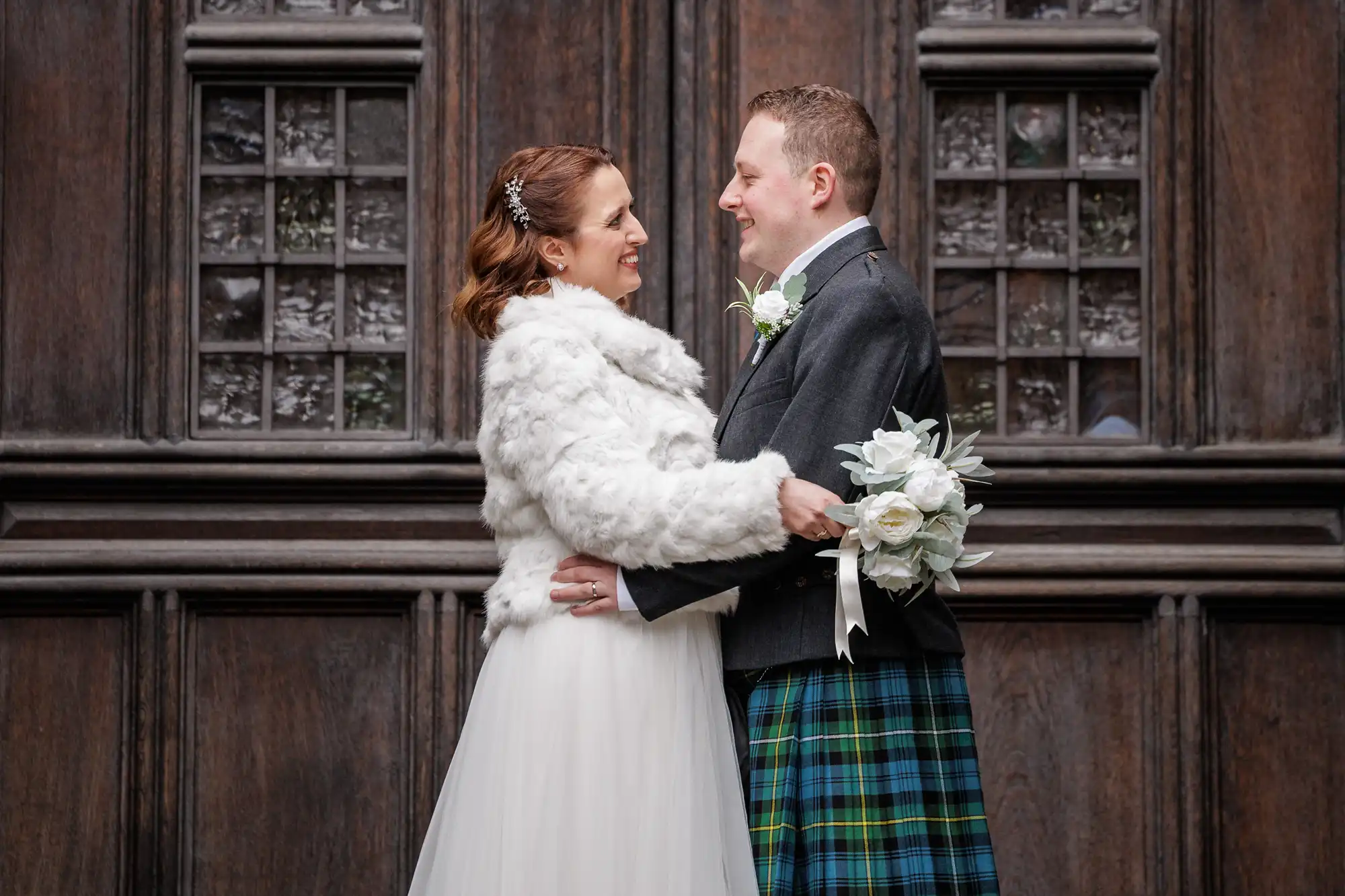 A bride in a white dress and faux fur jacket smiles at a groom wearing a kilt and jacket. They embrace and face each other, holding a bouquet of white flowers, standing in front of a large, ornate wooden door with glass panes.